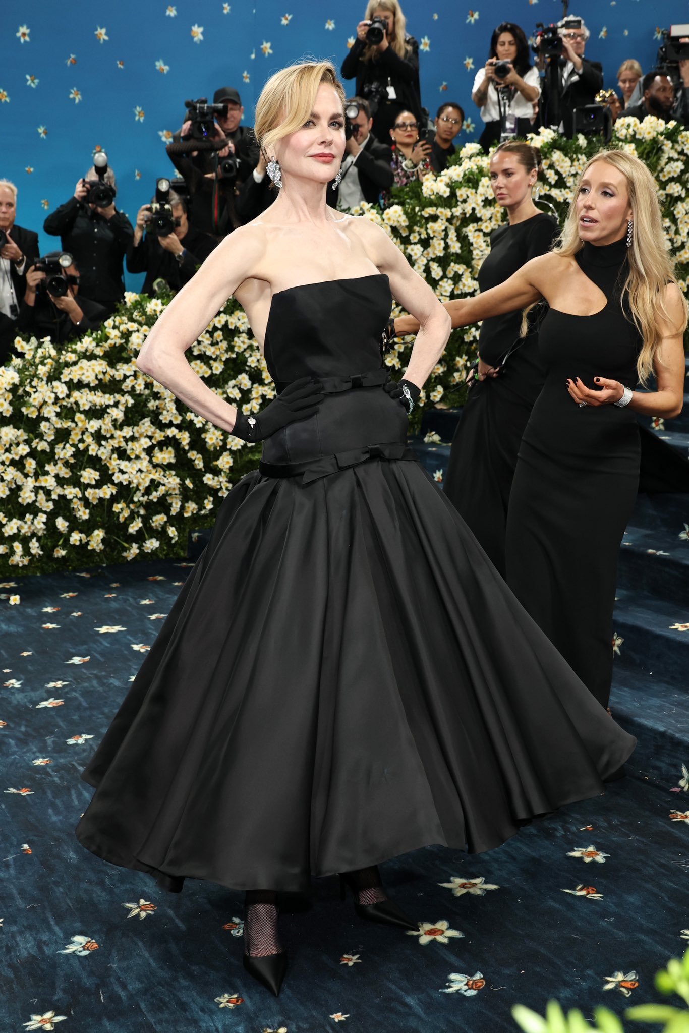 Celebridade em evento com vestido preto elegante no Met Gala 2025, cercada por fotógrafos e decoração floral.