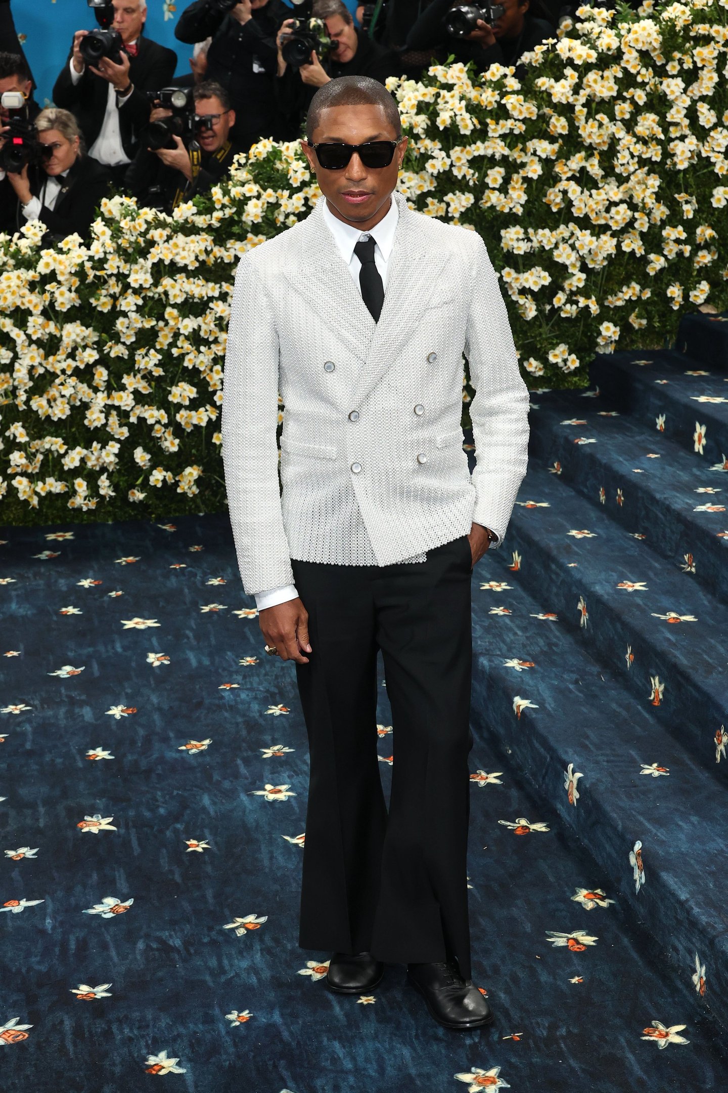Pessoa com terno branco e óculos escuros em evento do Met Gala 2025, rodeado por flores e fotógrafos.