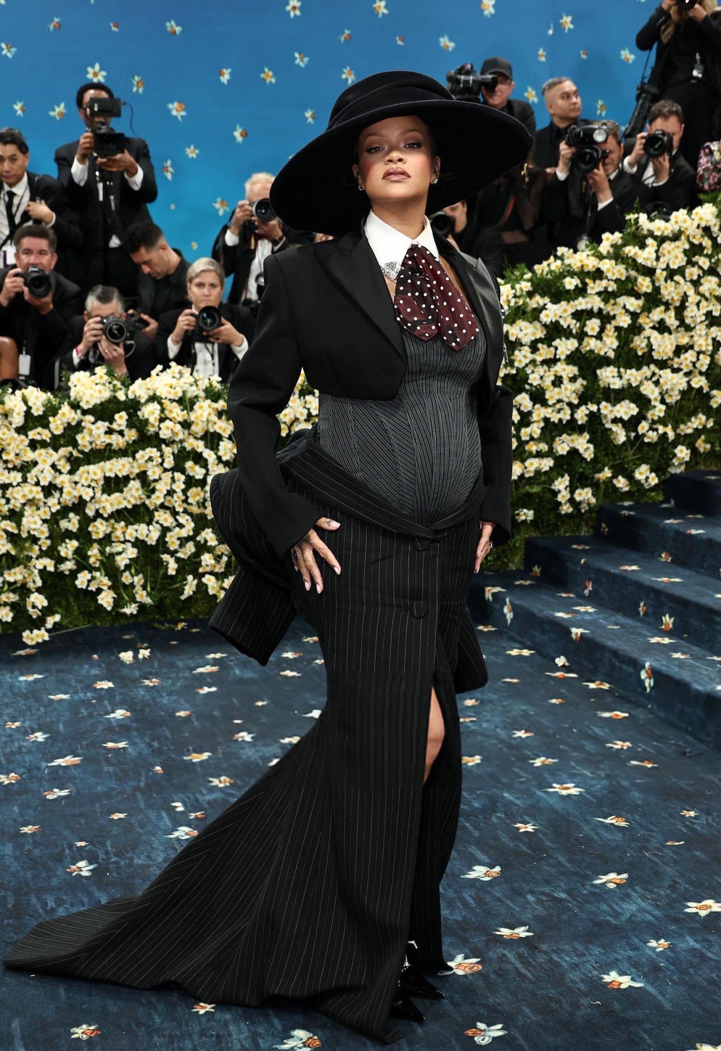 Pessoa usando traje risca de giz preto e chapéu largo no tapete com flores e fundo azul decorado.