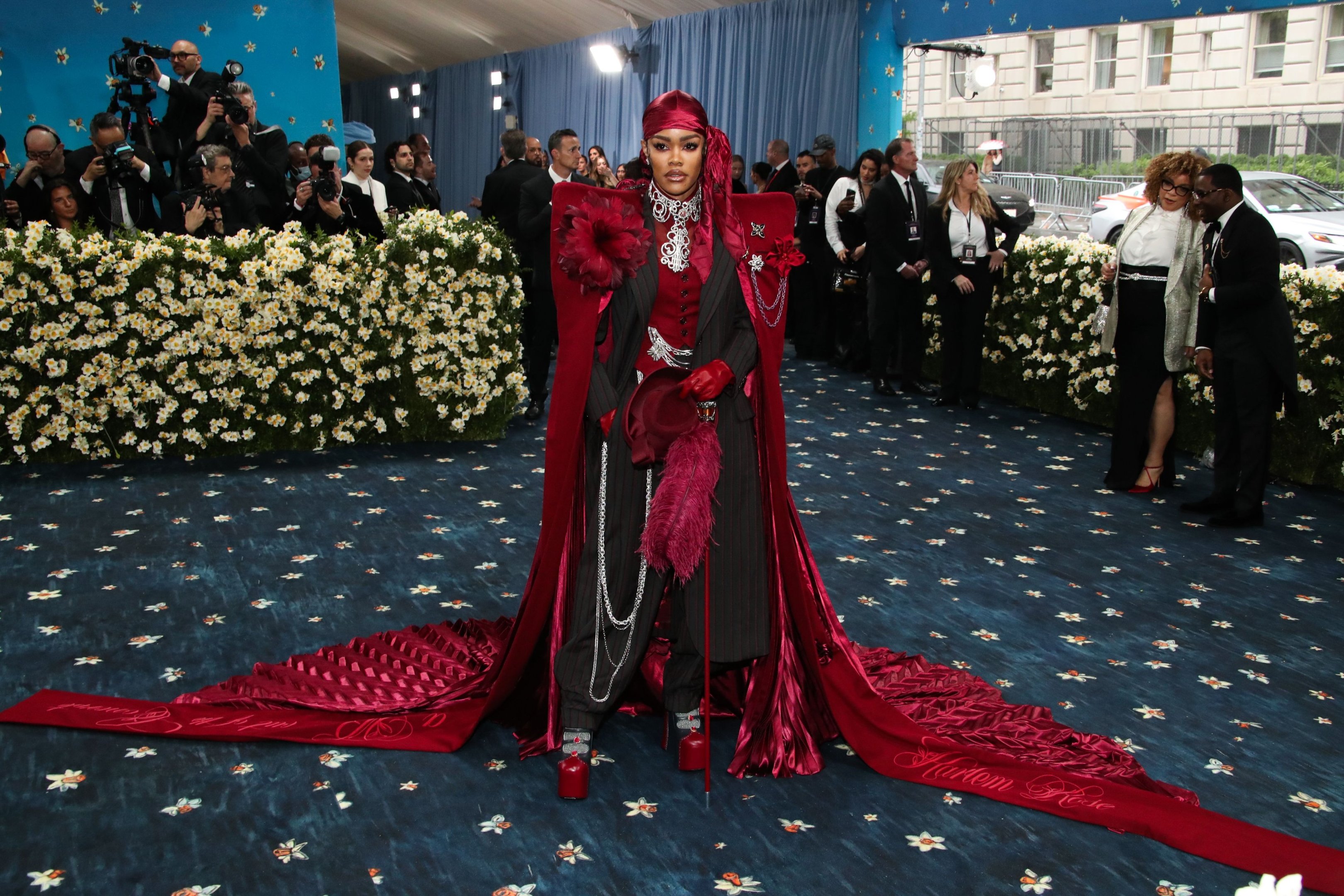 Celebridade em roupa vermelha marcante no Met Gala 2025, cercada por fotógrafos e decoração floral.