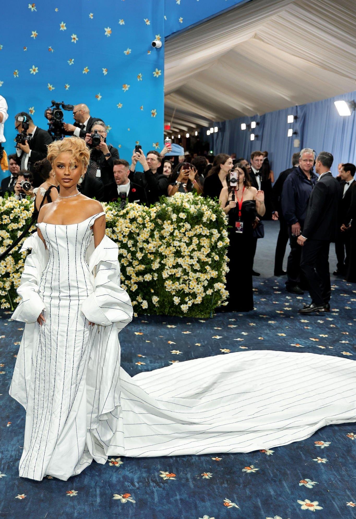 Celebridade em tapete azul com decorações florais, usando vestido branco com risca de giz e longa cauda.