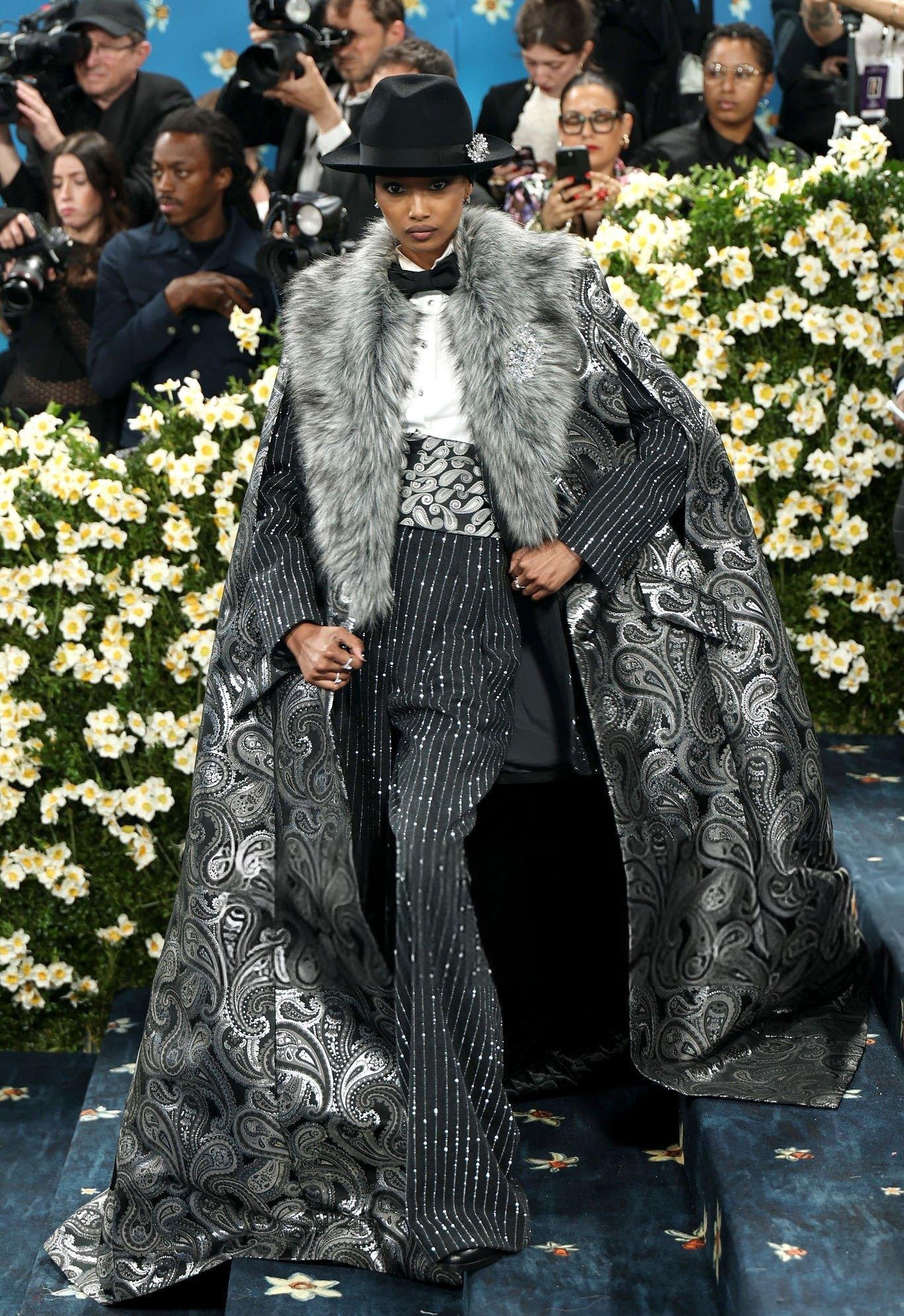 Pessoa elegante com capa luxuosa e terno risca de giz em evento badalado, cercada por fotógrafos e flores.