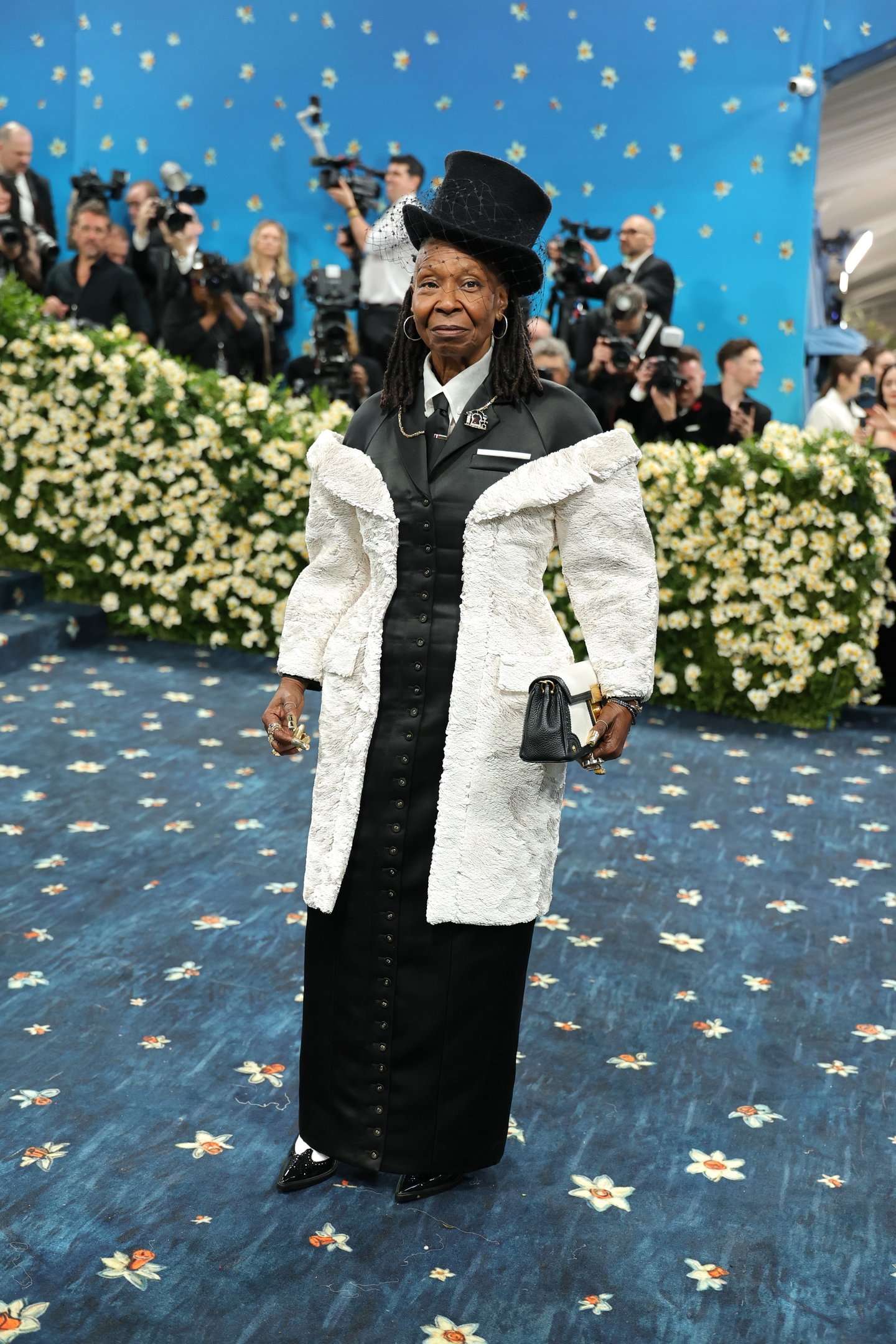 Pessoa vestida elegantemente com tema clássico durante o Met Gala 2025. Ao fundo, fotógrafos e decoração floral.