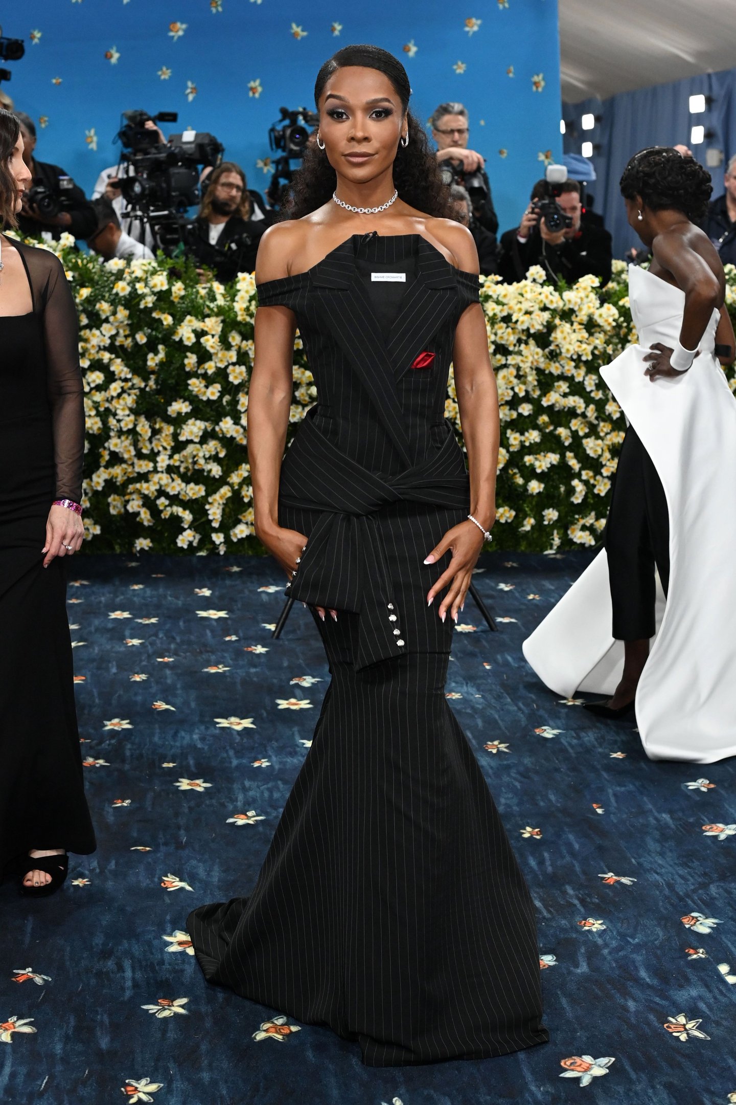 Mulher usando vestido preto com risca de giz em evento, com fundo floral e pessoas ao redor.