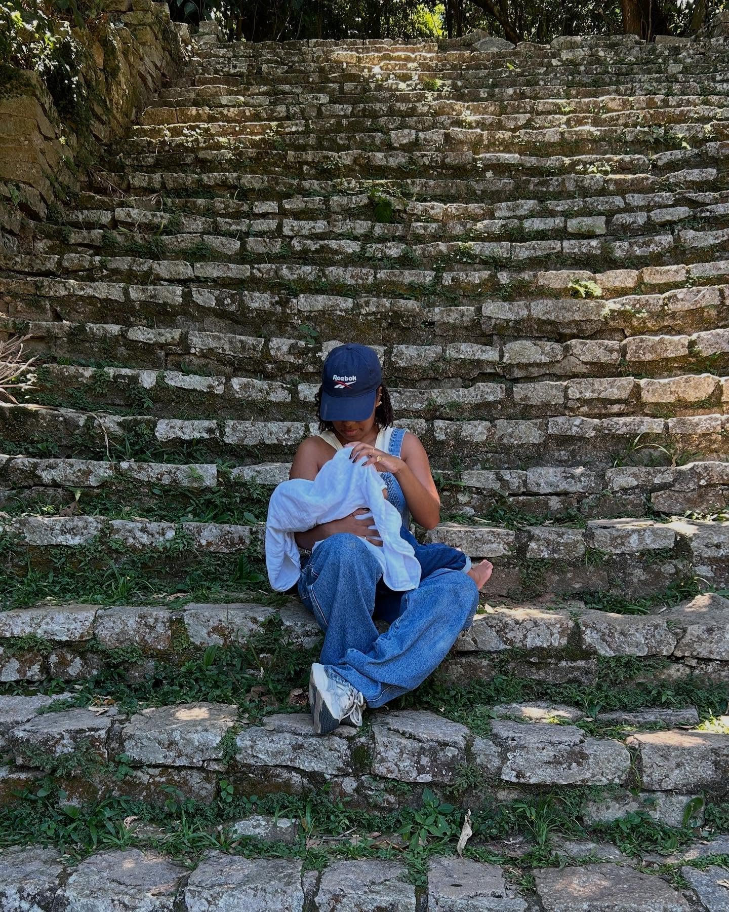 Mulher sentada em escadaria de pedra, segurando um bebê, simbolizando maternidade em ambiente natural.