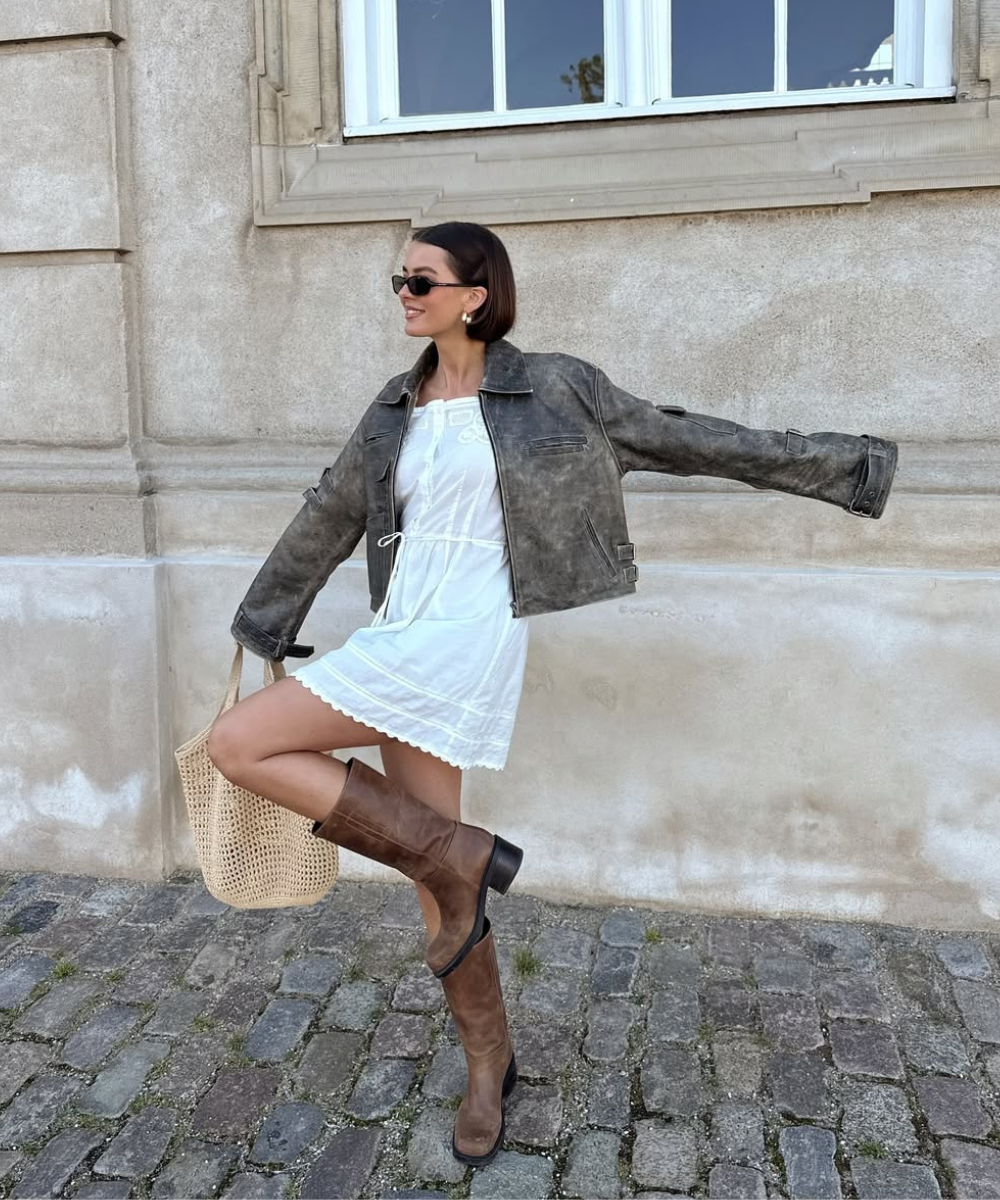 Mulher com mini dress branco, jaqueta cinza, botas marrons e bolsa de palha, posa alegremente em calçada de pedra.