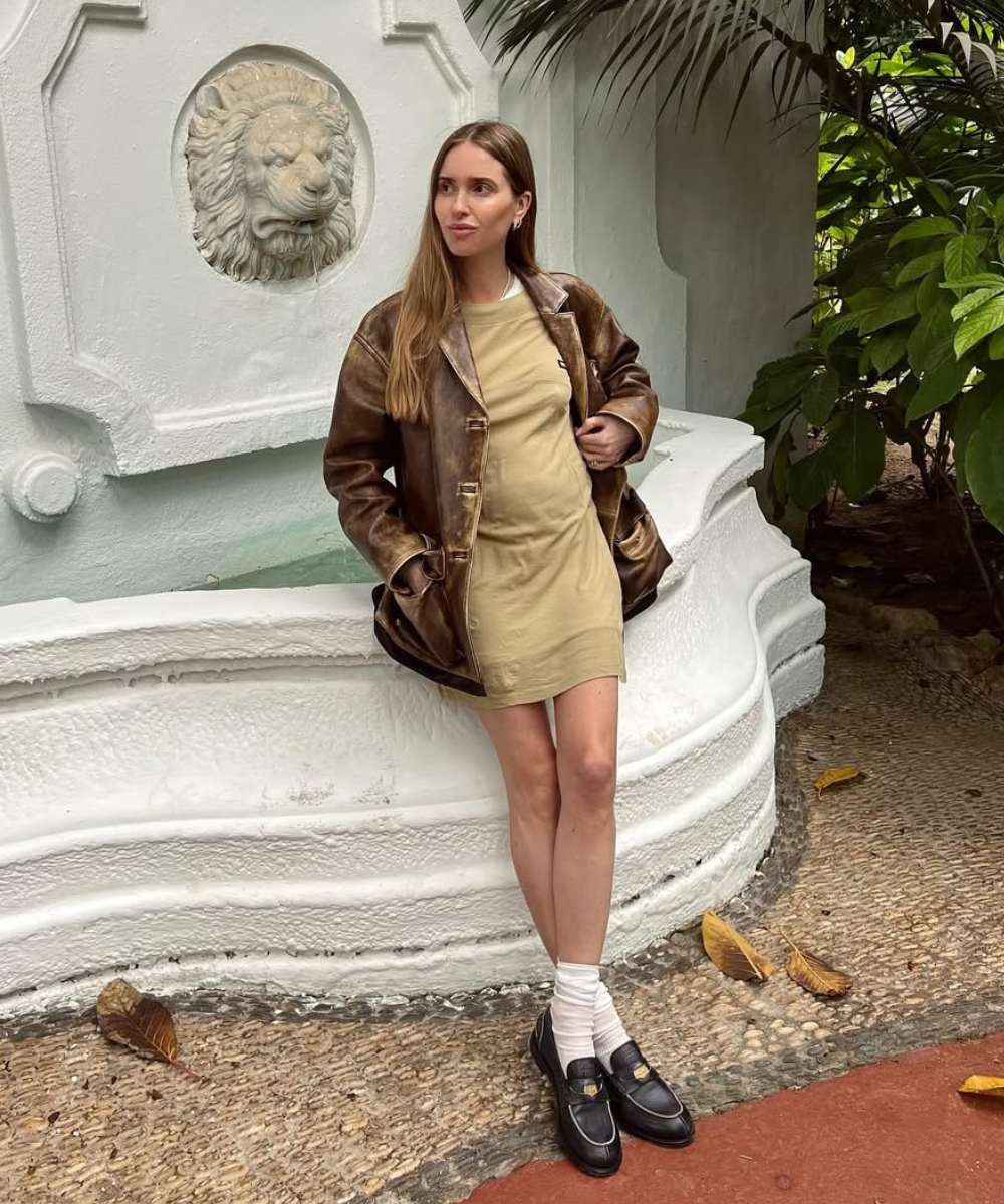 Mulher de mini dress bege e jaqueta marrom em pé, perto de fonte branca com escultura de leão.