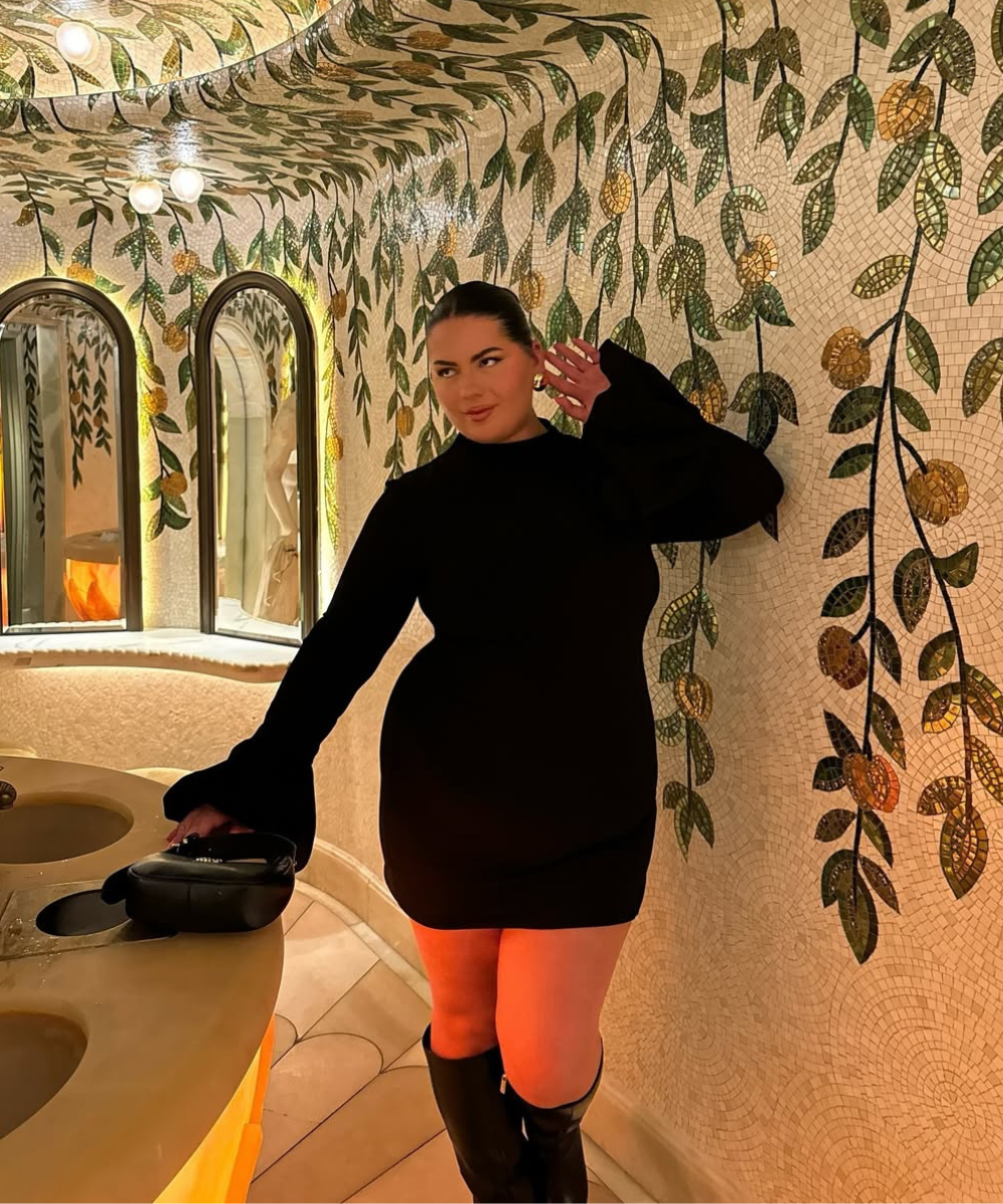 Mulher de mini vestido preto posa em banheiro decorado com mosaicos de folhas e flores.