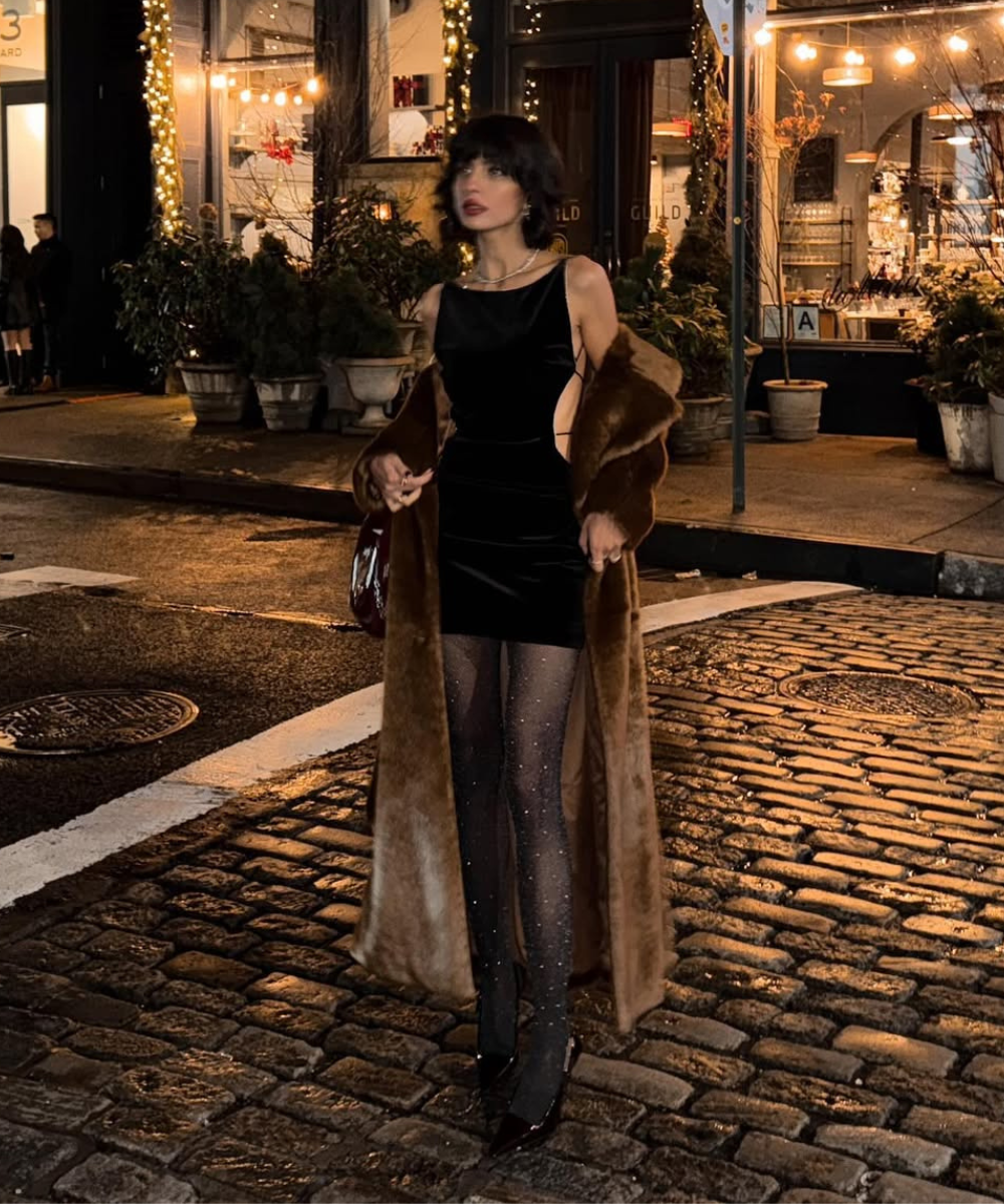 Pessoa com vestido curto preto e casaco de pele em rua iluminada por luzes festivas à noite.