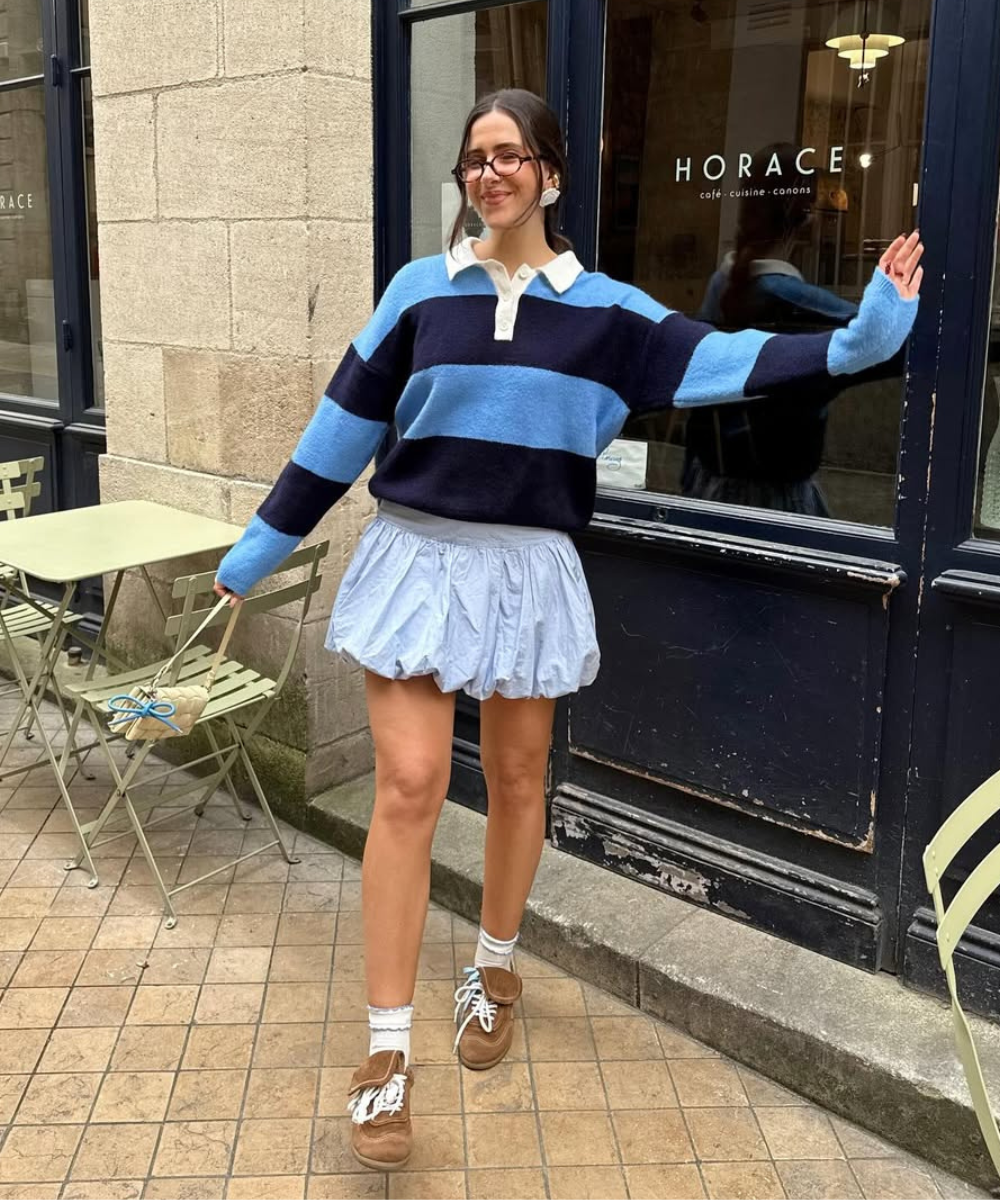 Pessoa sorrindo usa suéter listrado azul e preto sobre mini dress azul claro, em frente a uma cafeteria.