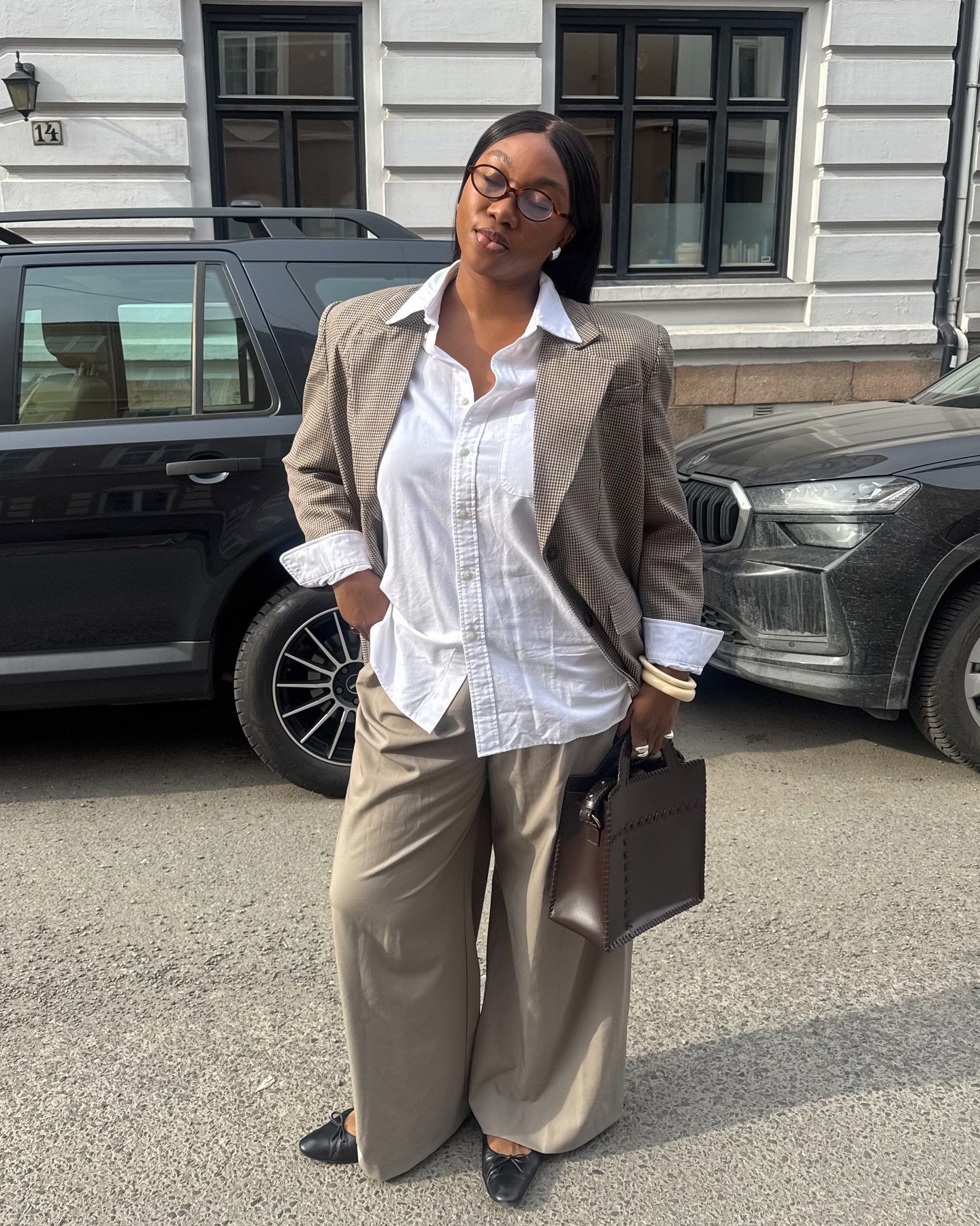 Mulher em moda executiva: blazer xadrez, camisa branca, calça larga bege, em frente a um carro preto.