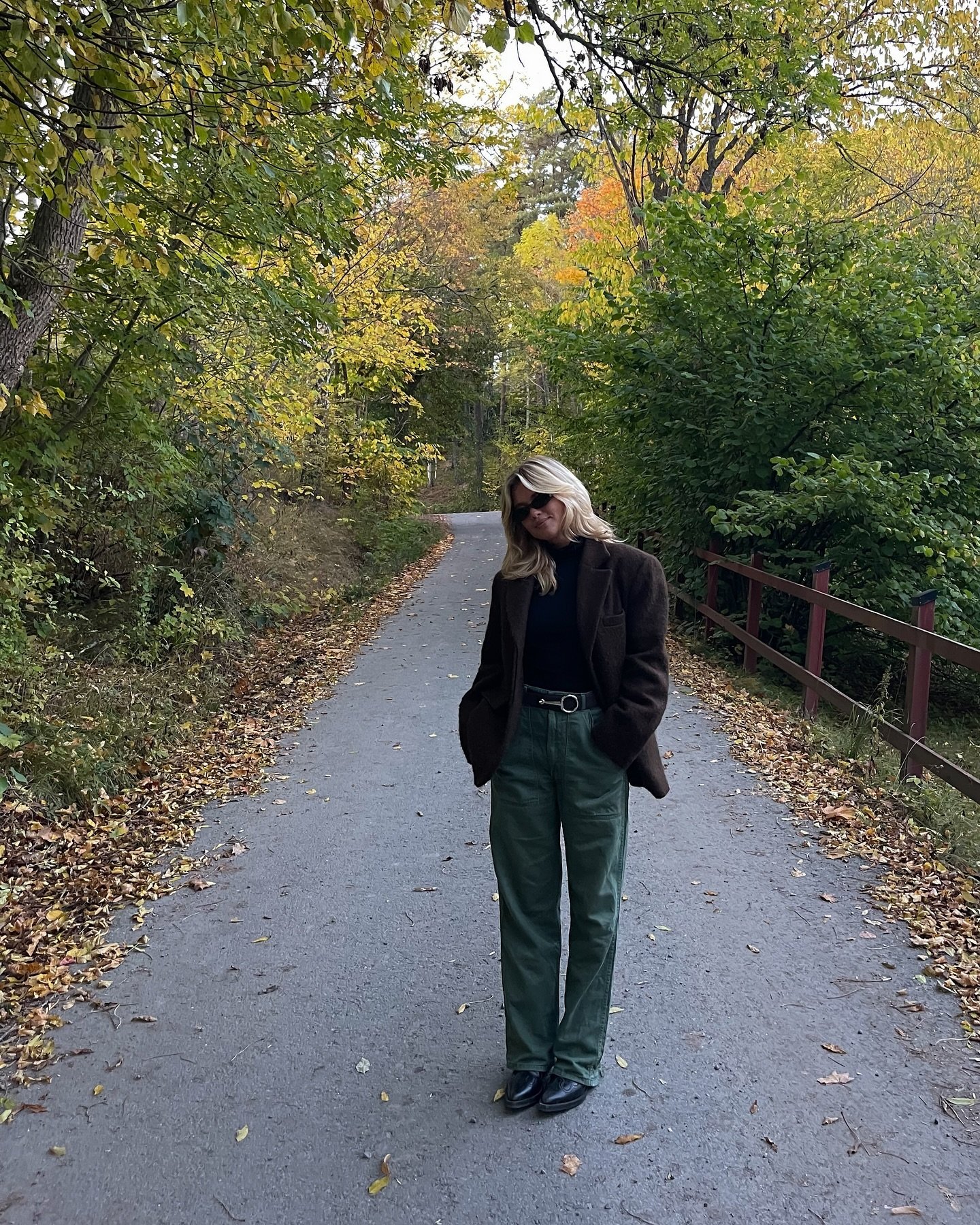 Mulher em roupa casual chique, com blazer marrom e calça verde, caminha por trilha arborizada no outono.