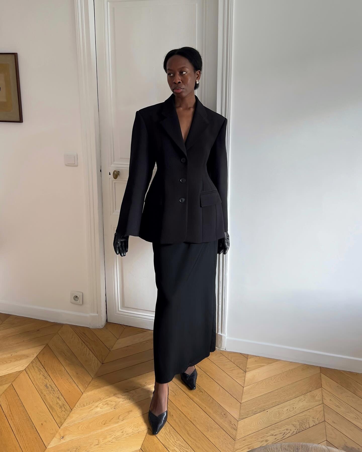 Vestindo traje elegante de moda executiva, mulher usa blazer preto, saia longa e luvas, em ambiente minimalista.