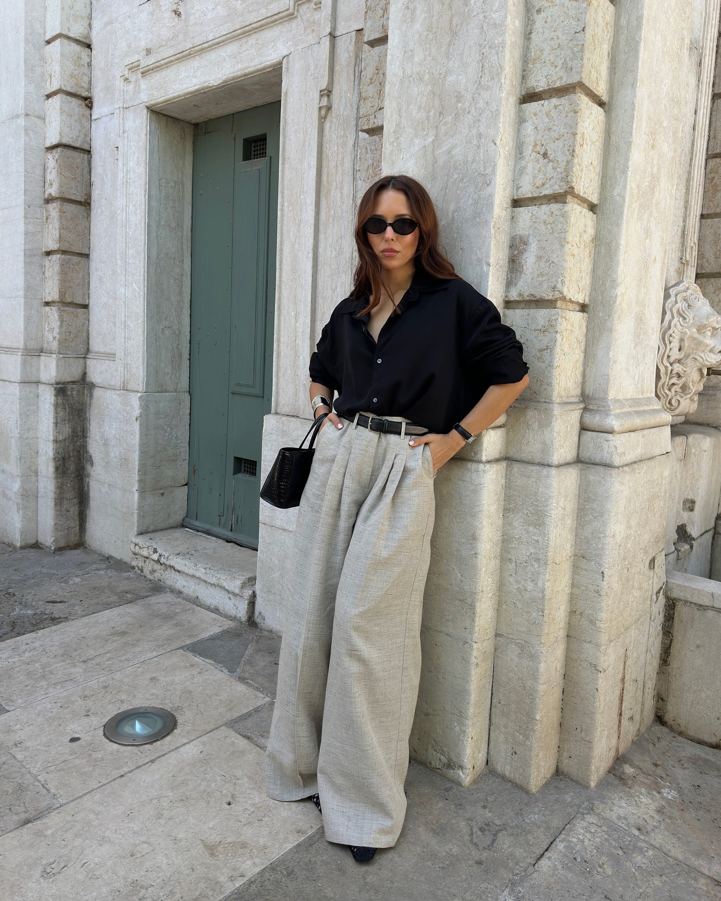 Pessoa usando moda executiva com calça larga cinza e camisa preta, óculos escuros e bolsa elegante, em ambiente urbano.