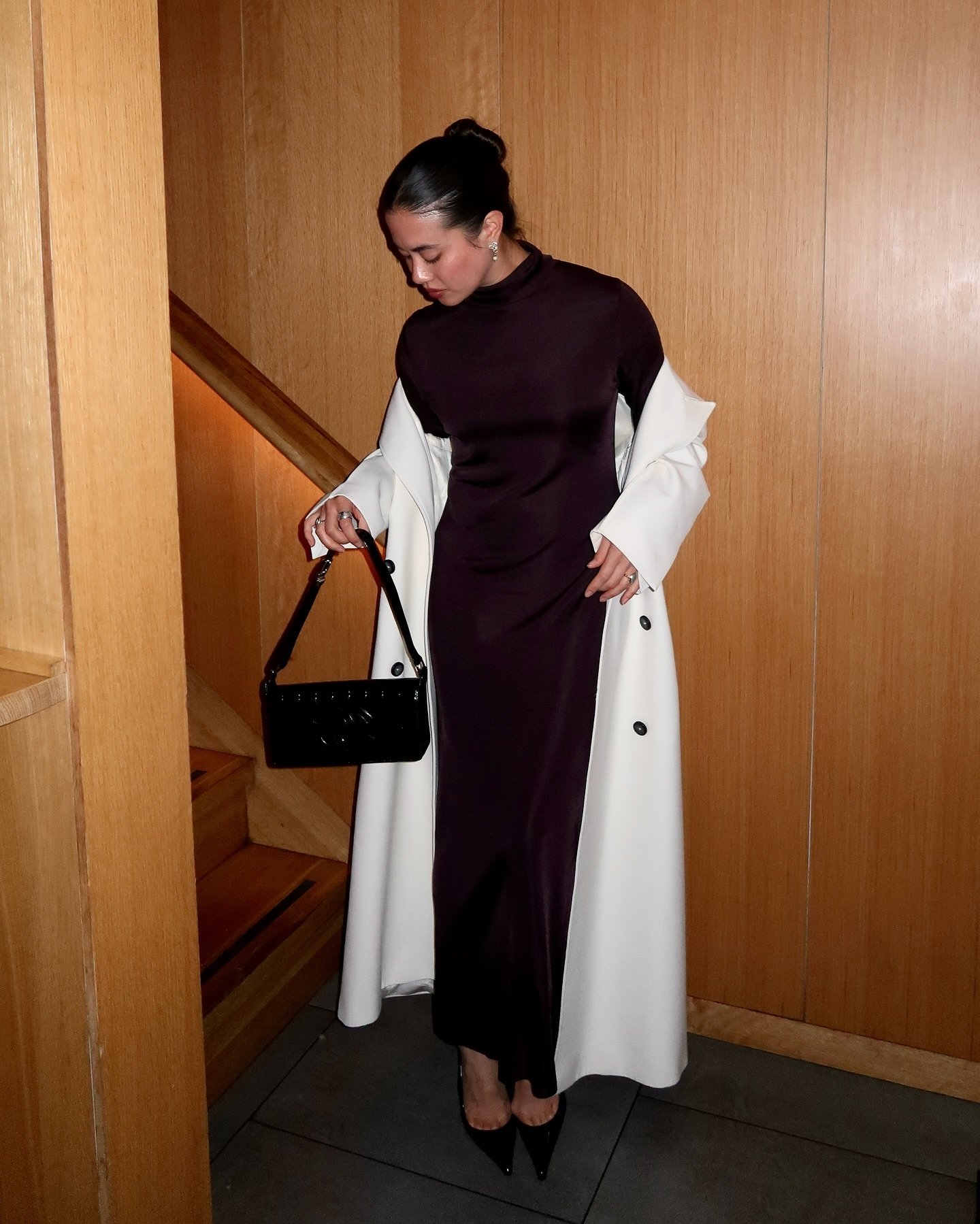 Mulher usa vestido longo e casaco branco elegante, segurando bolsa preta em cenário sofisticado, representando moda executiva.