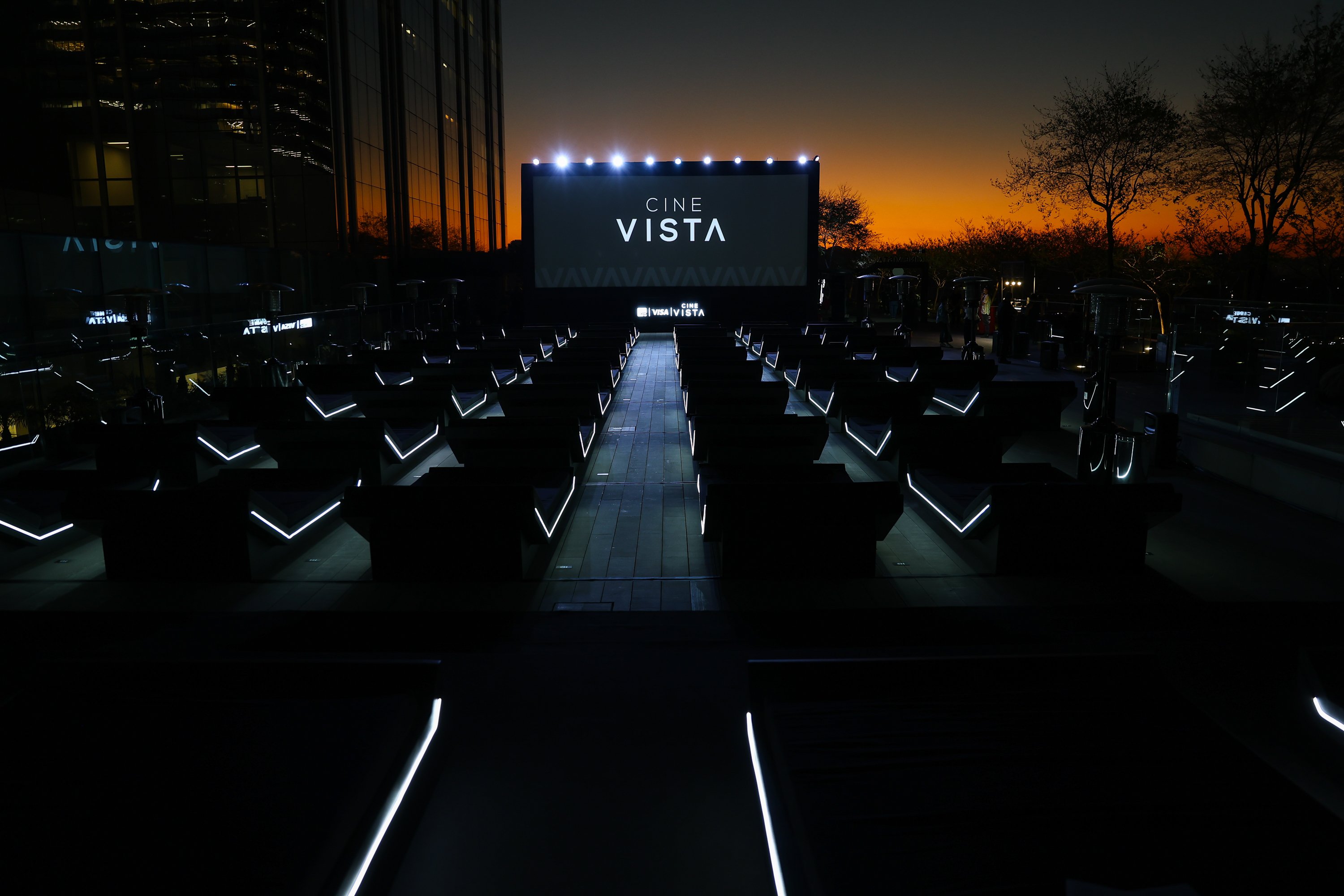 Tela de cinema ao ar livre do Cine Vista, com assentos iluminados ao anoitecer e um pôr do sol ao fundo.