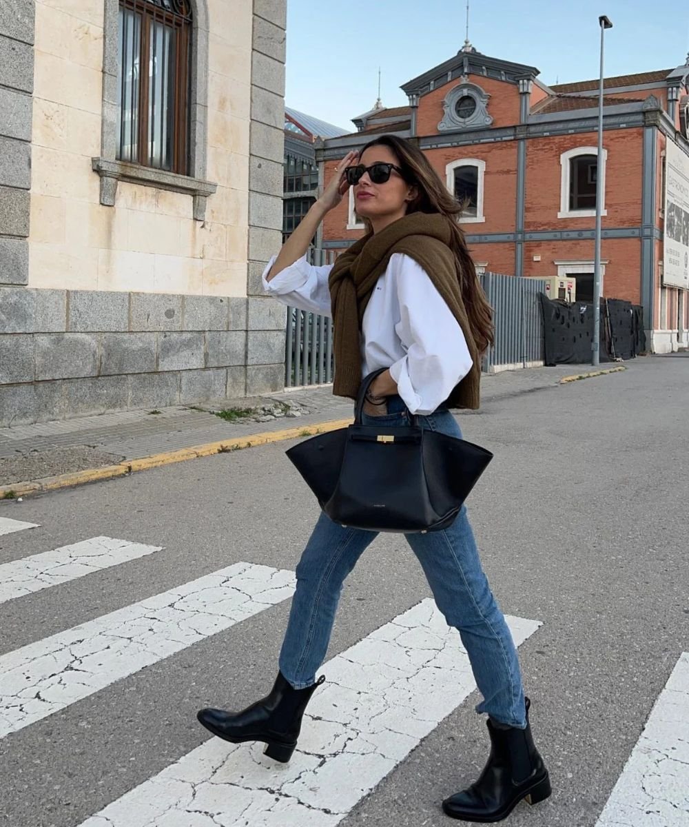 Pessoa atravessa rua usando look com bota cano curto, calça jeans e bolsa preta. Look estiloso com blusa branca e suéter marrom.