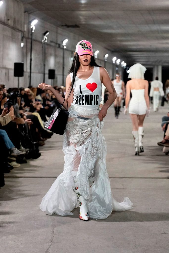 Modelo desfila em passarela usando top com estampa de coração e saia rendada longa, segurando bolsa e vestindo boné rosa. Na moda