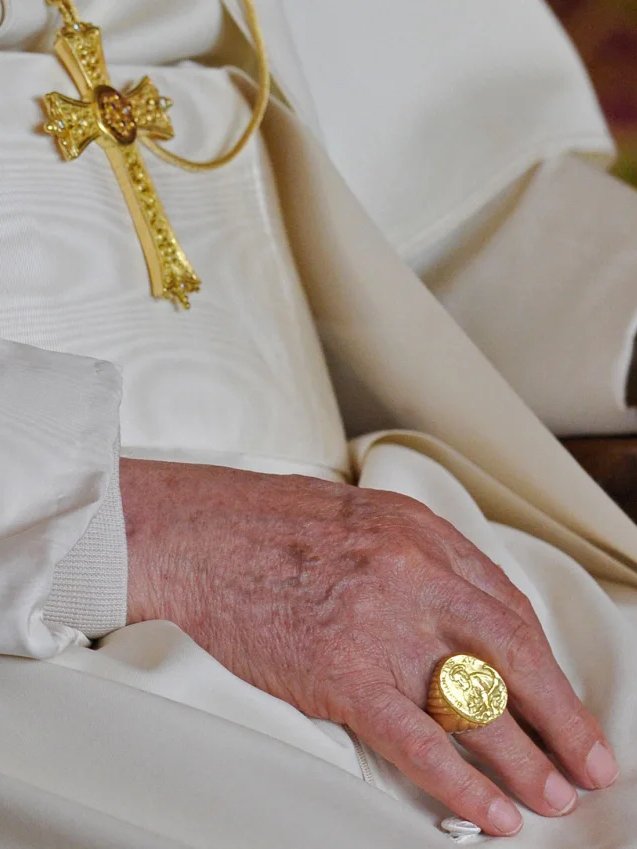 Mão com anel dourado sobre roupa branca. O anel simboliza legado e tradição na história dos papas.