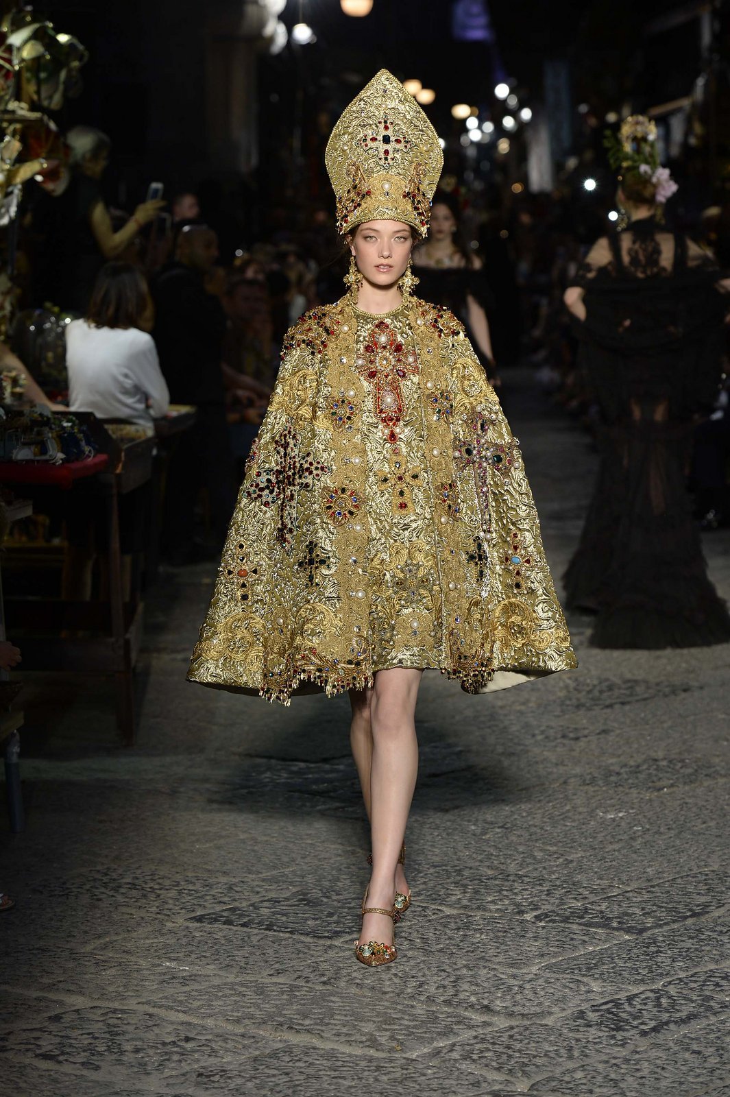 Modelo com traje dourado ornamentado e mitra papal, inspirando-se na história dos papas, em desfile noturno.