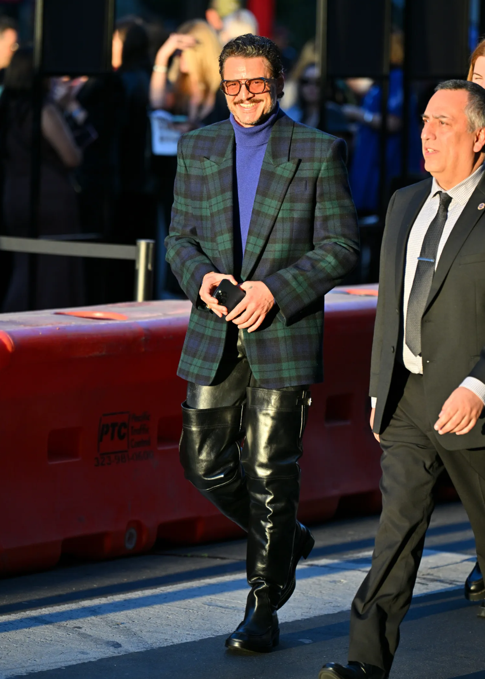 A imagem mostra Pedro Pascal sorrindo, usando casaco xadrez, suéter azul e botas altas.
