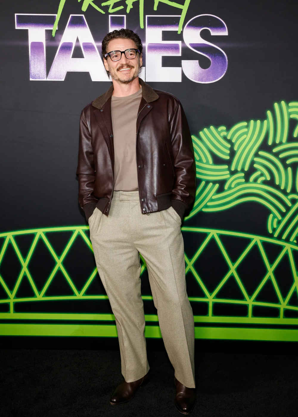 Um homem de óculos, jaqueta de couro marrom e calças bege, sorrindo em um evento com fundo colorido.