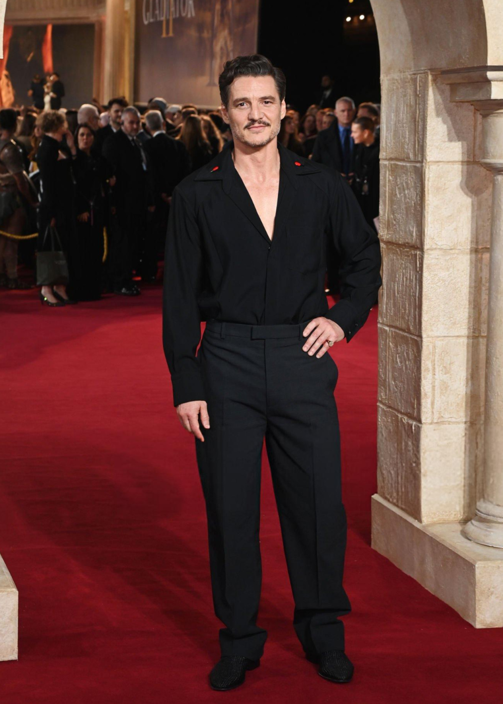 Um homem com cabelo curto em evento de gala usa camisa e calça pretas sobre tapete vermelho. Atrás, grupo de pessoas.