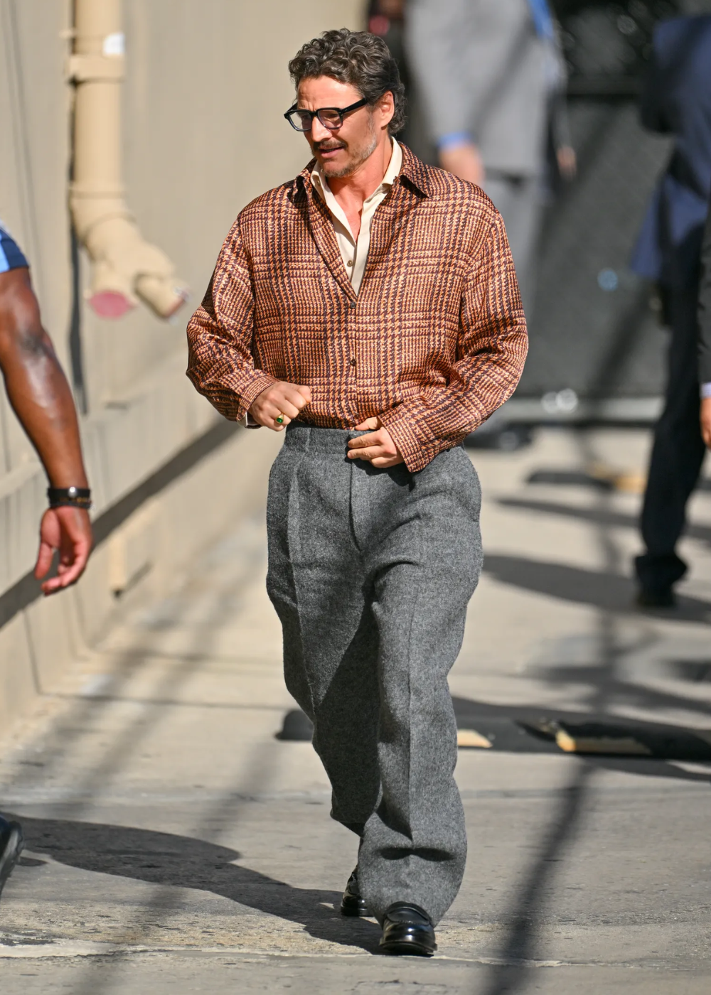 Pedro Pascal usando um look com camisa xadrez e calça cinza enquanto anda na rua.