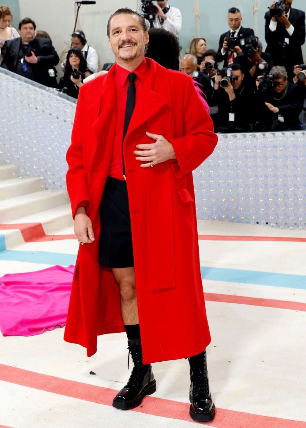 Pedro Pascal usando casaco vermelho e botas está posando em frente a fotógrafos.