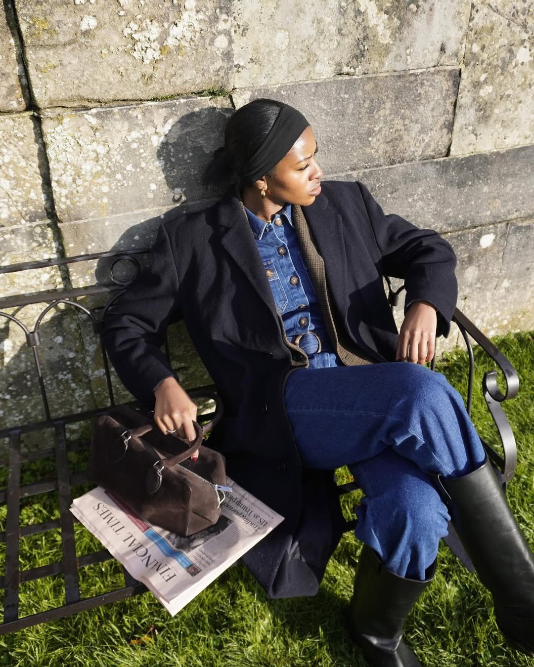 Mulher de cabelos presos, sentada em banco ao ar livre, usando jeans e jaqueta escura. Jornal e bolsa ao lado.