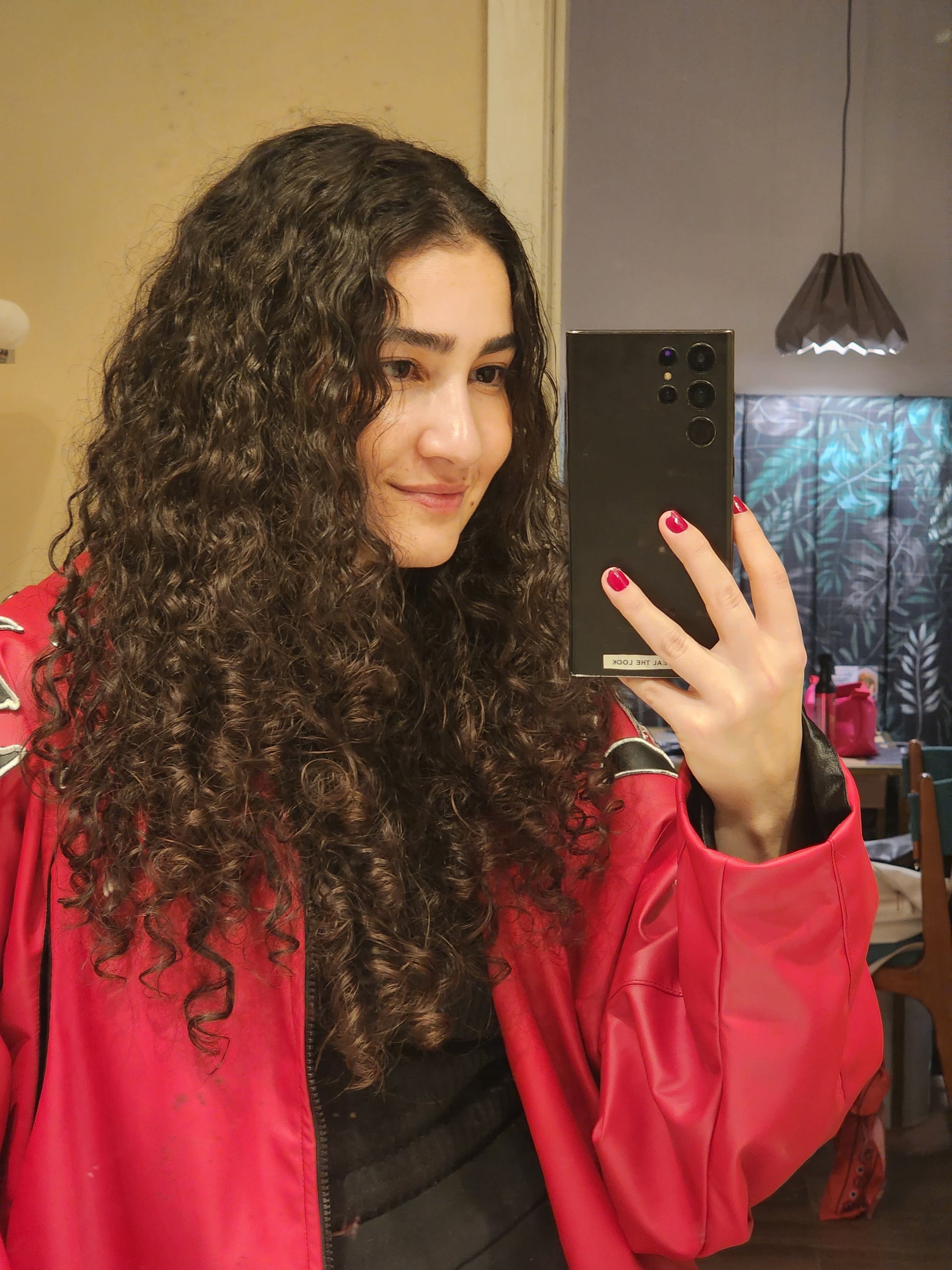 Mulher com cabelos cacheados e brilhantes, vestindo jaqueta vermelha, tirando selfie. Cabelo bem cuidado com efeito antifrizz.