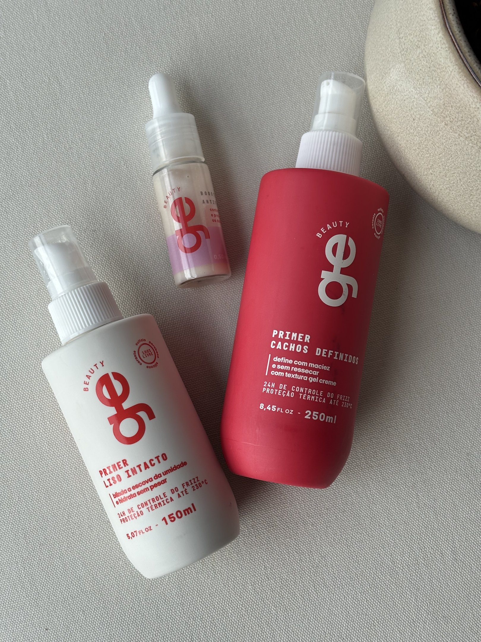 Três frascos de produtos capilares para primer, incluindo um para cachos e outro anti-frizz, sobre superfície clara. Primer de cabelo