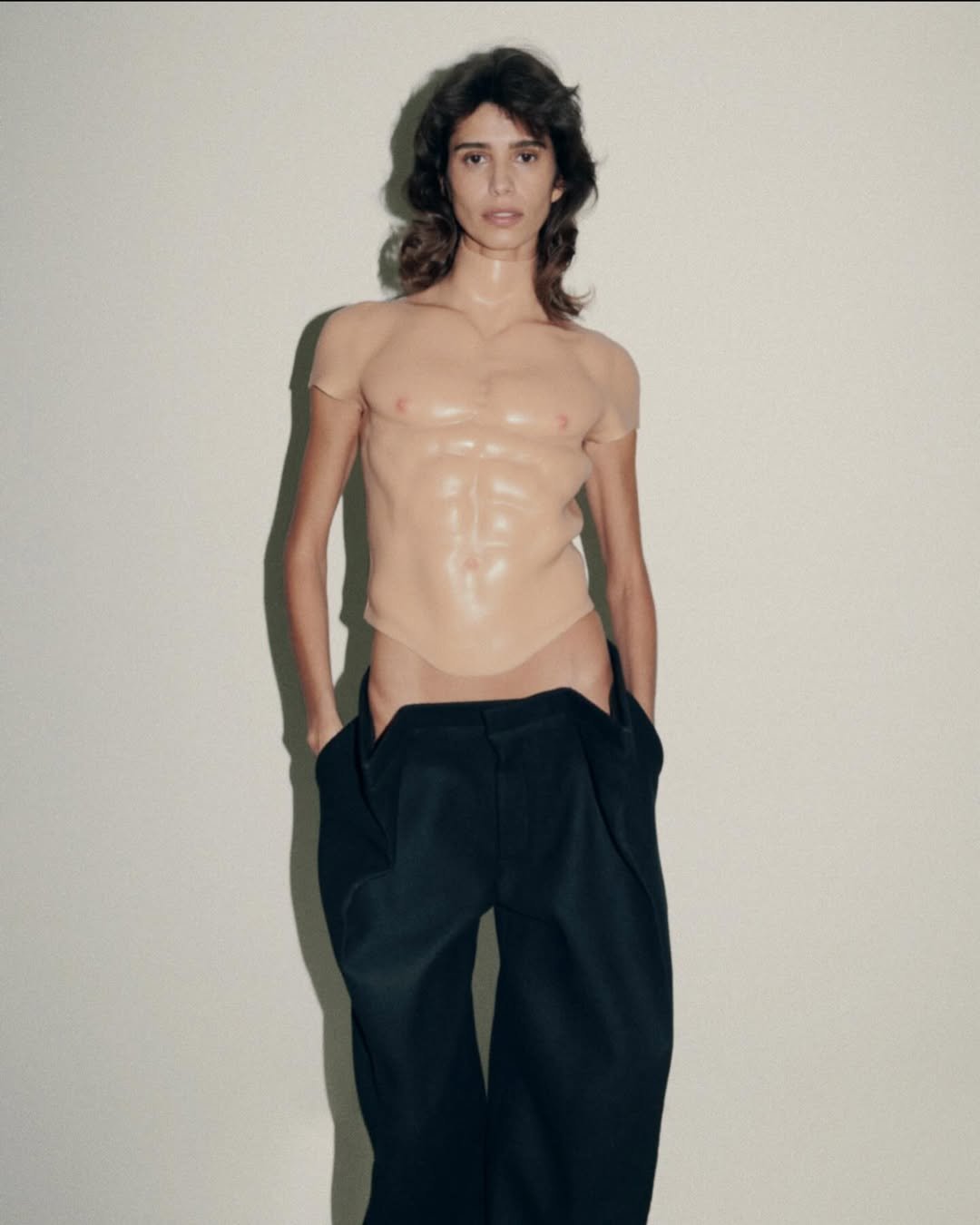 Pessoa vestindo uma prótese de musculatura no tronco e calças largas escuras, criando um visual artístico e moderno. Protein chic