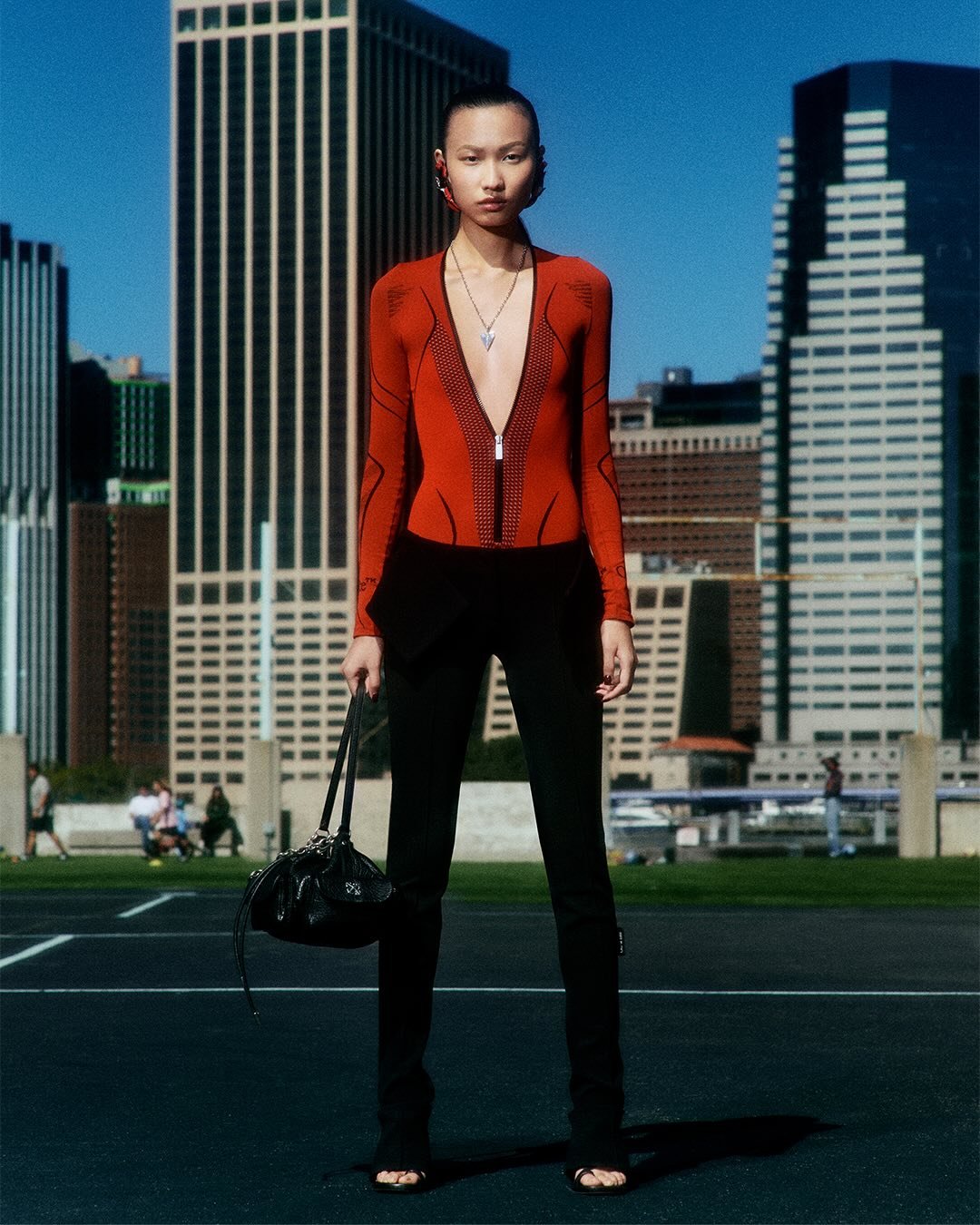 Pessoa com roupa moderna em frente a prédios altos, segurando uma bolsa preta. Estilo elegante e sofisticado. Protein chic