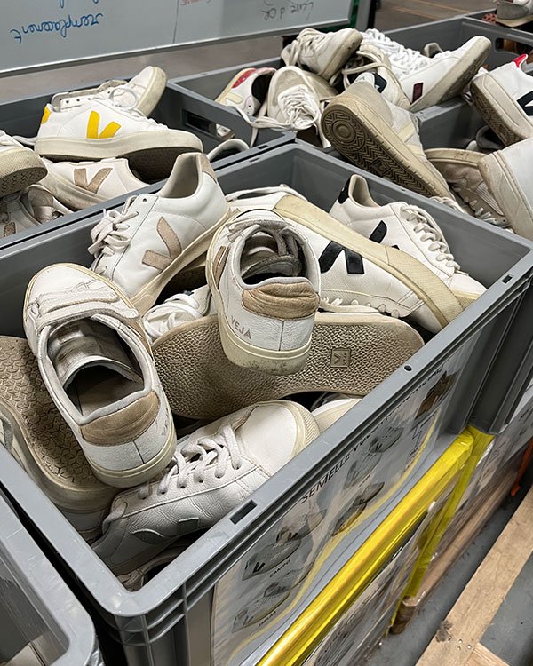 Tênis brancos e bege da marca VEJA empilhados em caixas cinzas sobre prateleiras.