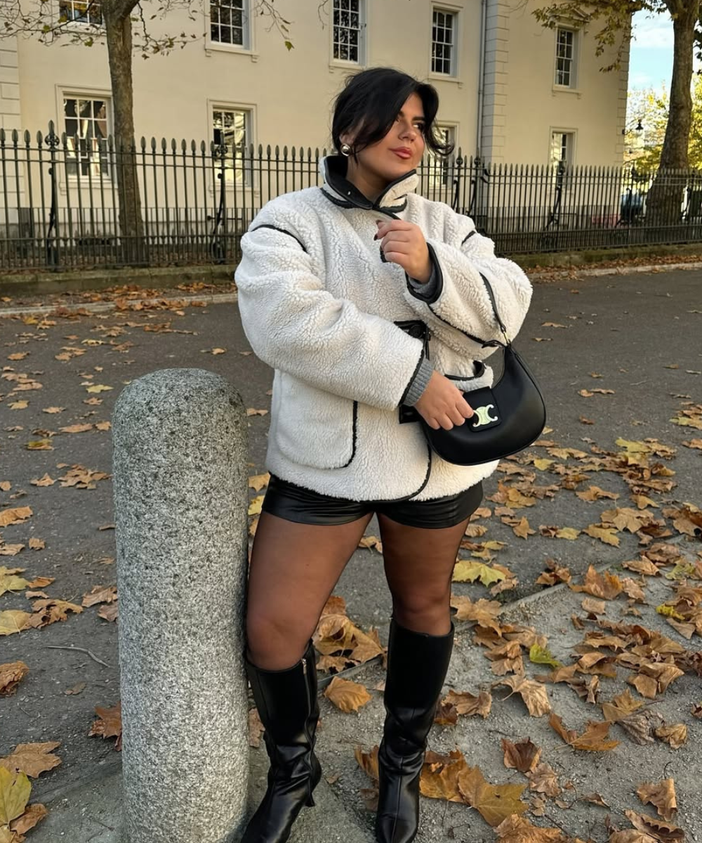 Pessoa com bolsa e shorts de couro fake em ambiente urbano, rodeada por folhas secas.