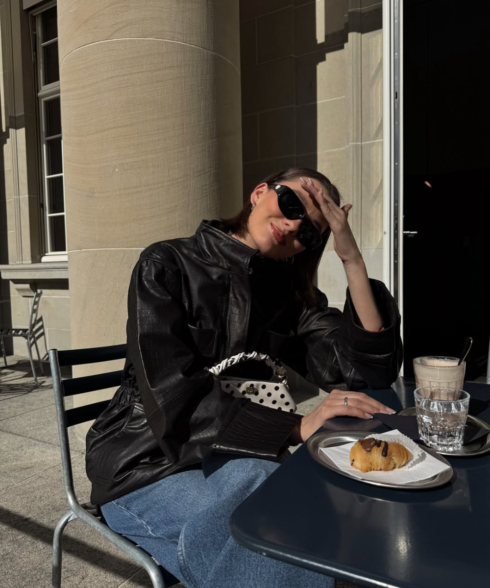 Pessoa com roupa de couro fake sentada em uma mesa ao ar livre, com croissant e bebidas.