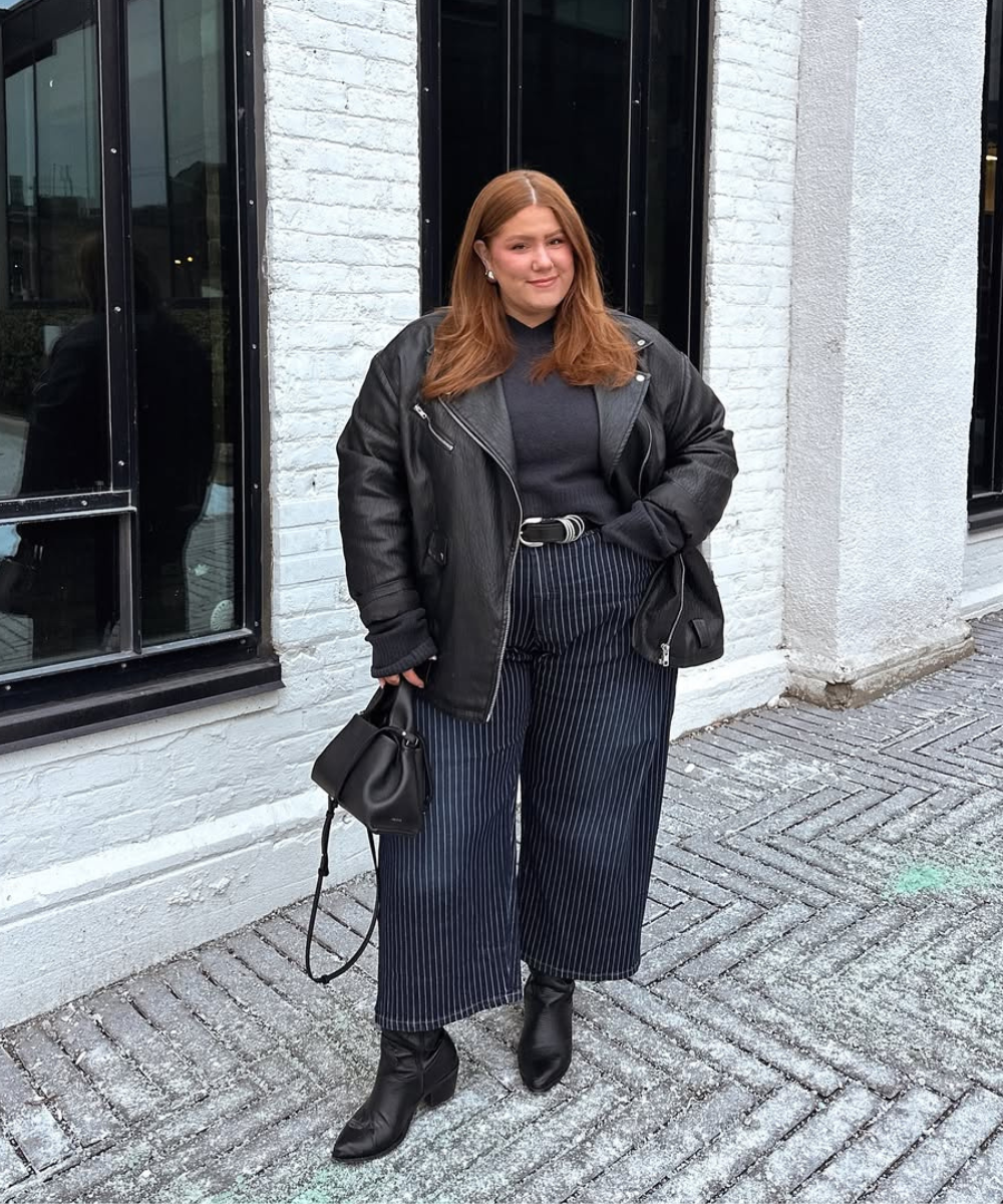 Pessoa com jaqueta de couro fake, blusa escura, calça listrada e botas pretas. Carrega bolsa pequena.