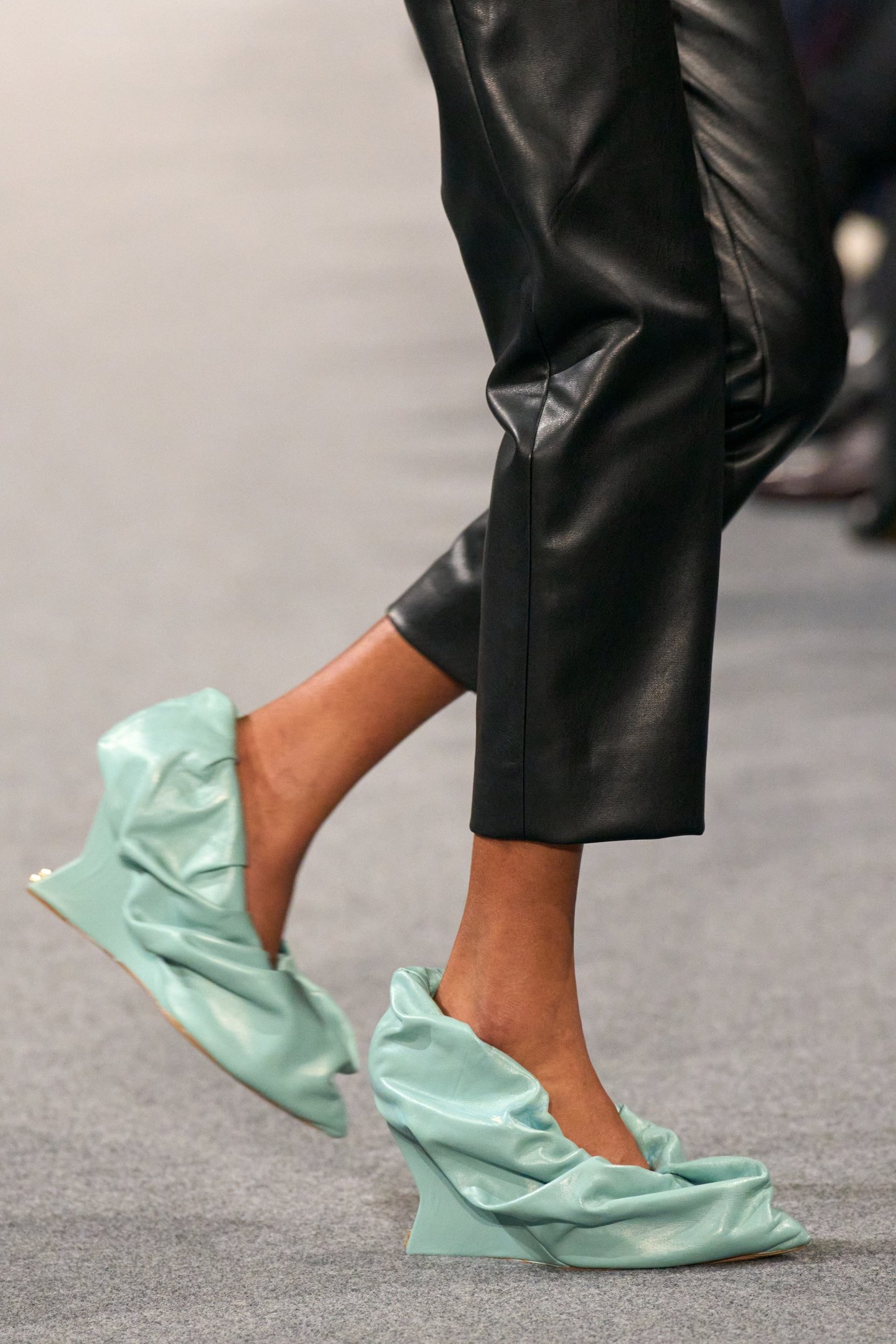 Modelo usando sapatos verde-claros com salto anabela, calça preta de couro, em desfile.