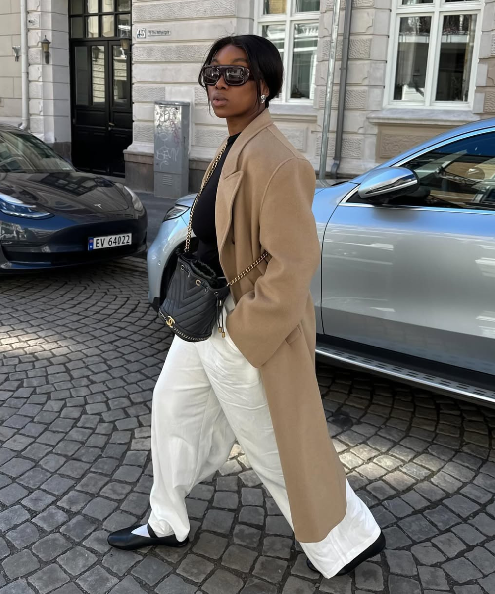 Pessoa caminhando na calçada de casaco bege, calça branca e sapatilha elegante preta.