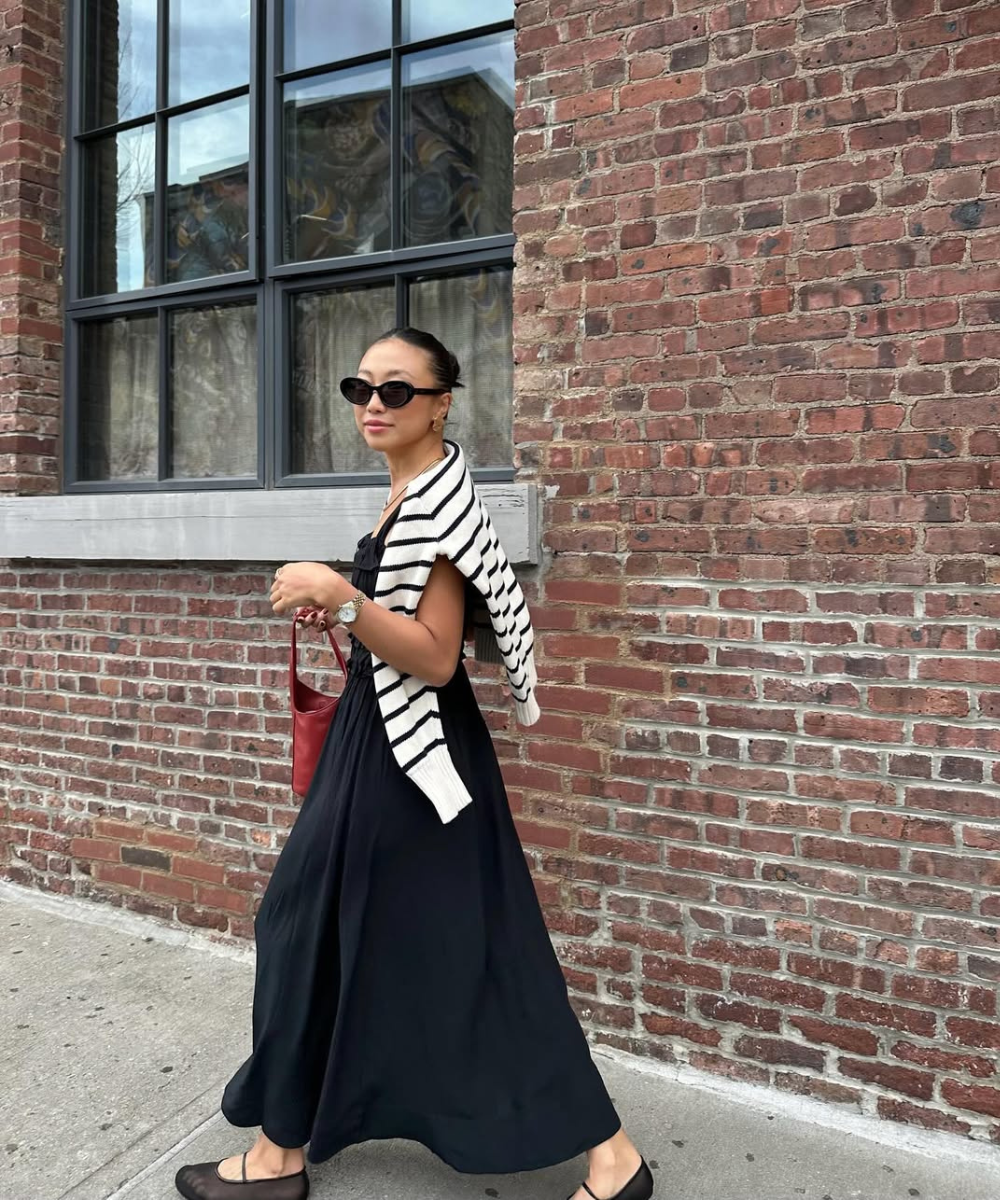 Pessoa de vestido preto, usando óculos escuros, caminha em frente a uma parede de tijolos, com sapatilhas elegantes.