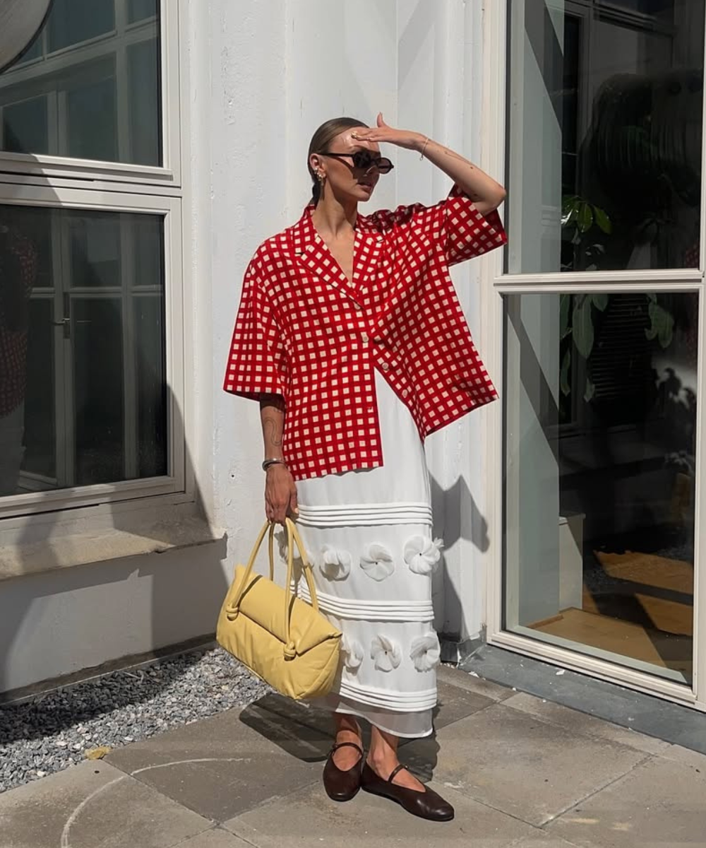 Mulher veste camisa vermelha quadriculada, saia branca longa e sapatilha elegante marrom. Carrega bolsa amarela.