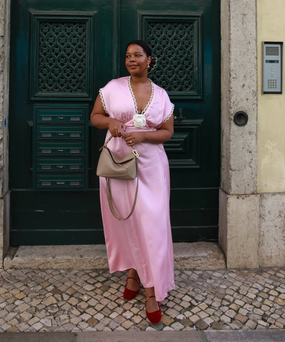 Mulher em frente a porta verde, usando vestido rosa e sapatilhas elegantes vermelhas, segurando bolsa bege.