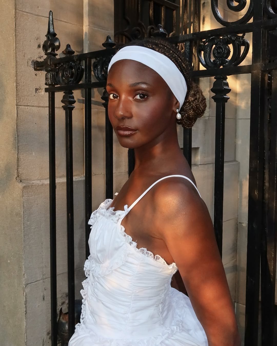 Pessoa em vestido branco e tiara posa com confiança, ressaltando tendência de beleza clássica.