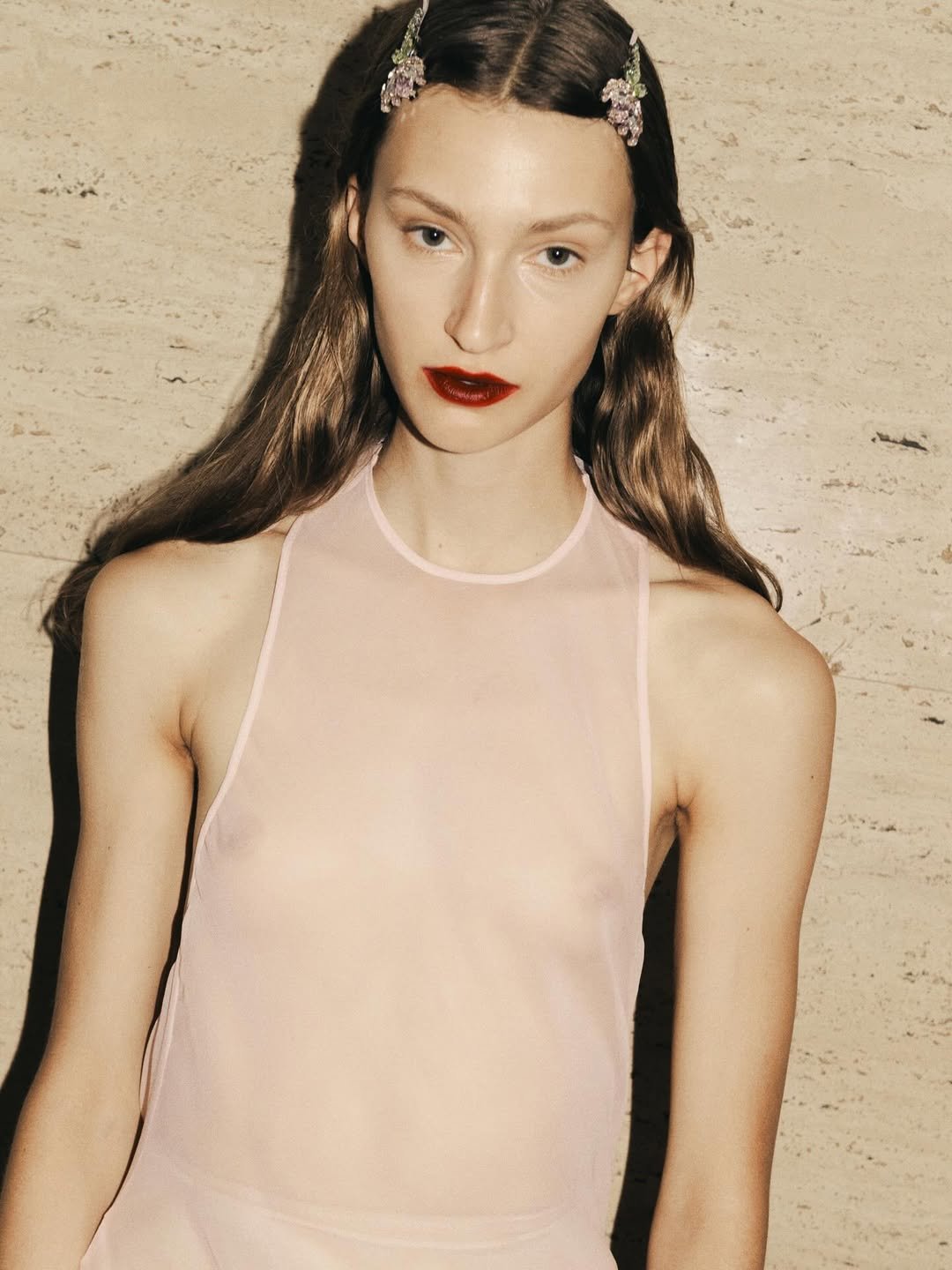 Mulher com cabelo ondulado, batom vermelho e presilhas florais, veste blusa transparente, refletindo tendência de beleza.