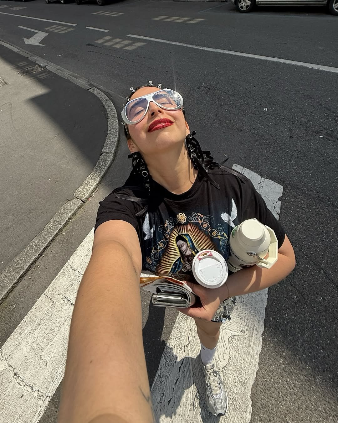 Pessoa sorridente segurando copo de café e acessório funcional - garrafa térmica, óculos grandes, na rua.