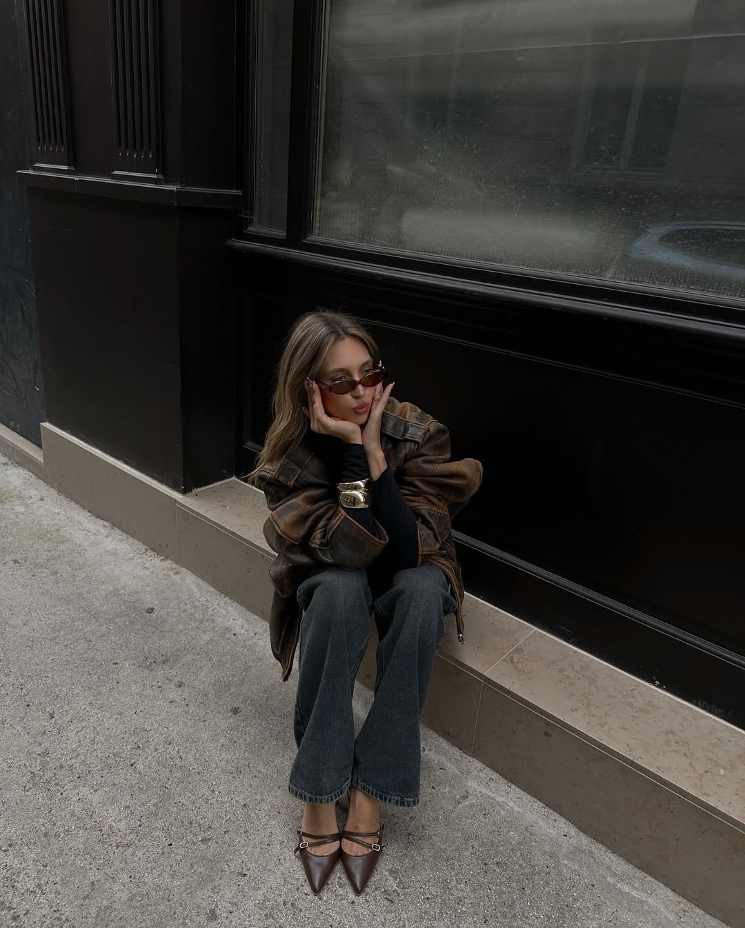 Mulher sentada em calçada, vestindo jaqueta  e scarpin marrom, óculos de sol e calça jeans.
