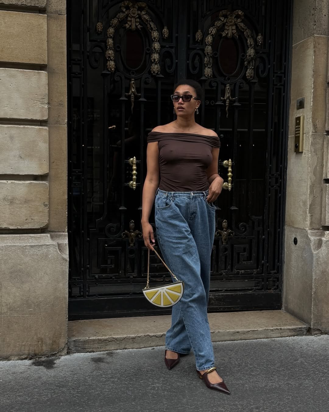 Pessoa com blusa marrom e calça jeans, segurando uma bolsa amarela em forma de limão, usa scarpin marrom, em frente a uma porta ornamentada.