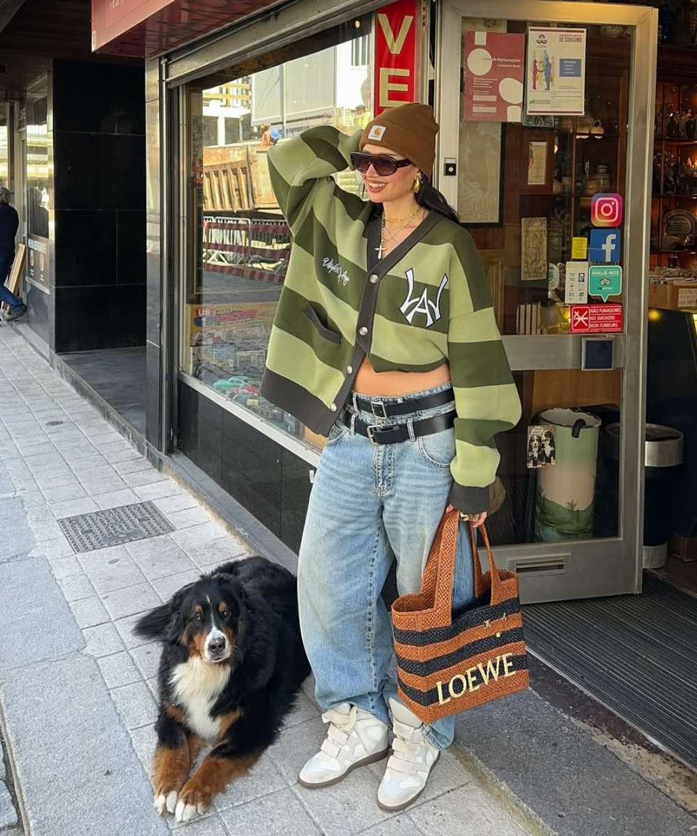 Mulher com suéter listrado verde, jeans largos e tênis, ao lado de cachorro, em frente a loja. 