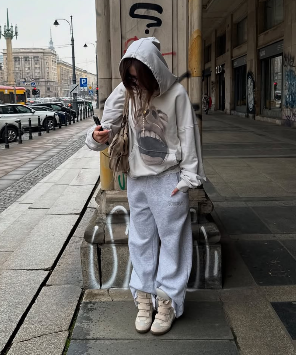Pessoa com moletom cinza e capuz, calça e tênis grossos em calçada urbana grafitada.